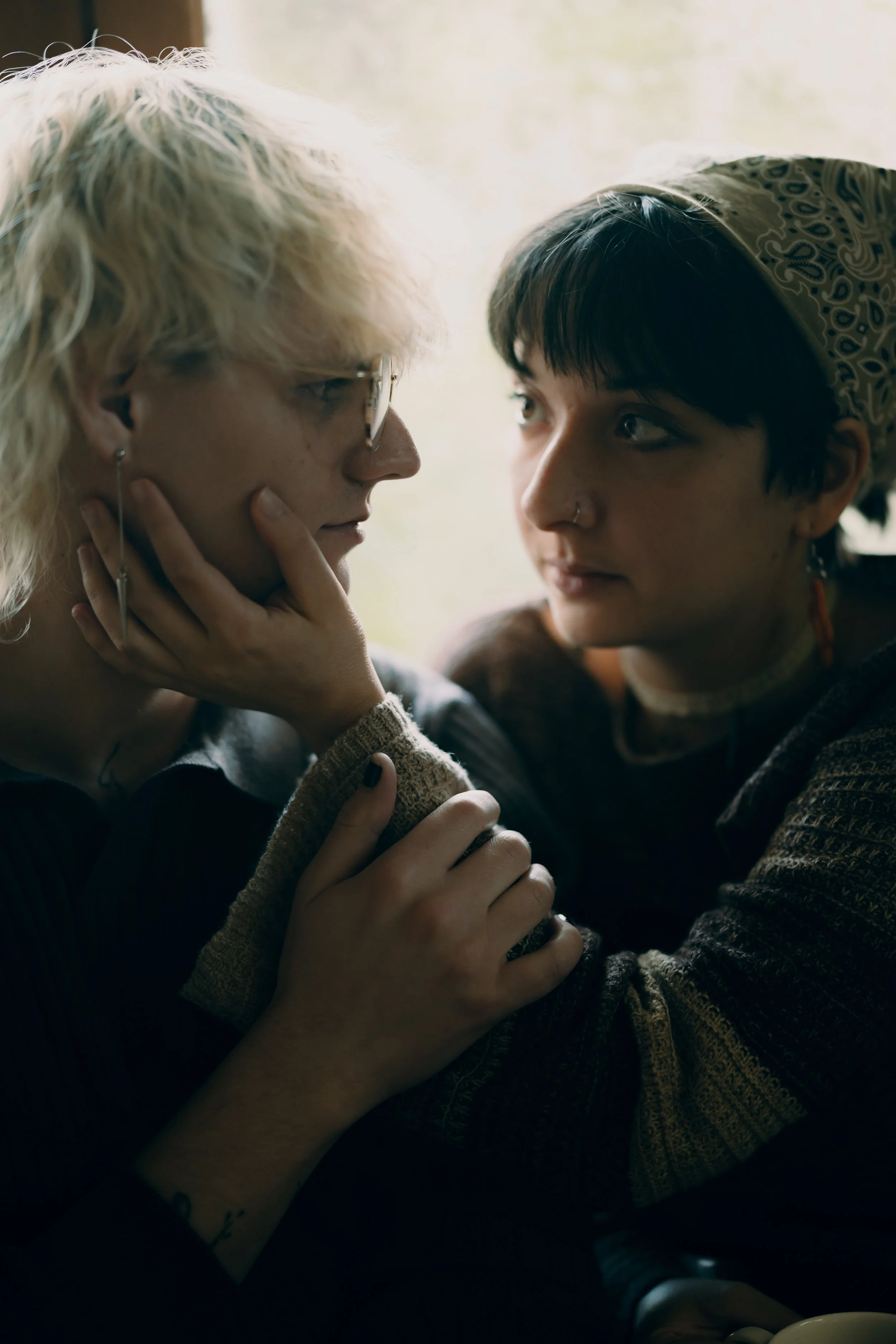 Two women face each other closely, one gently holding the other's face and hand, in an intimate moment inside a room with soft light.