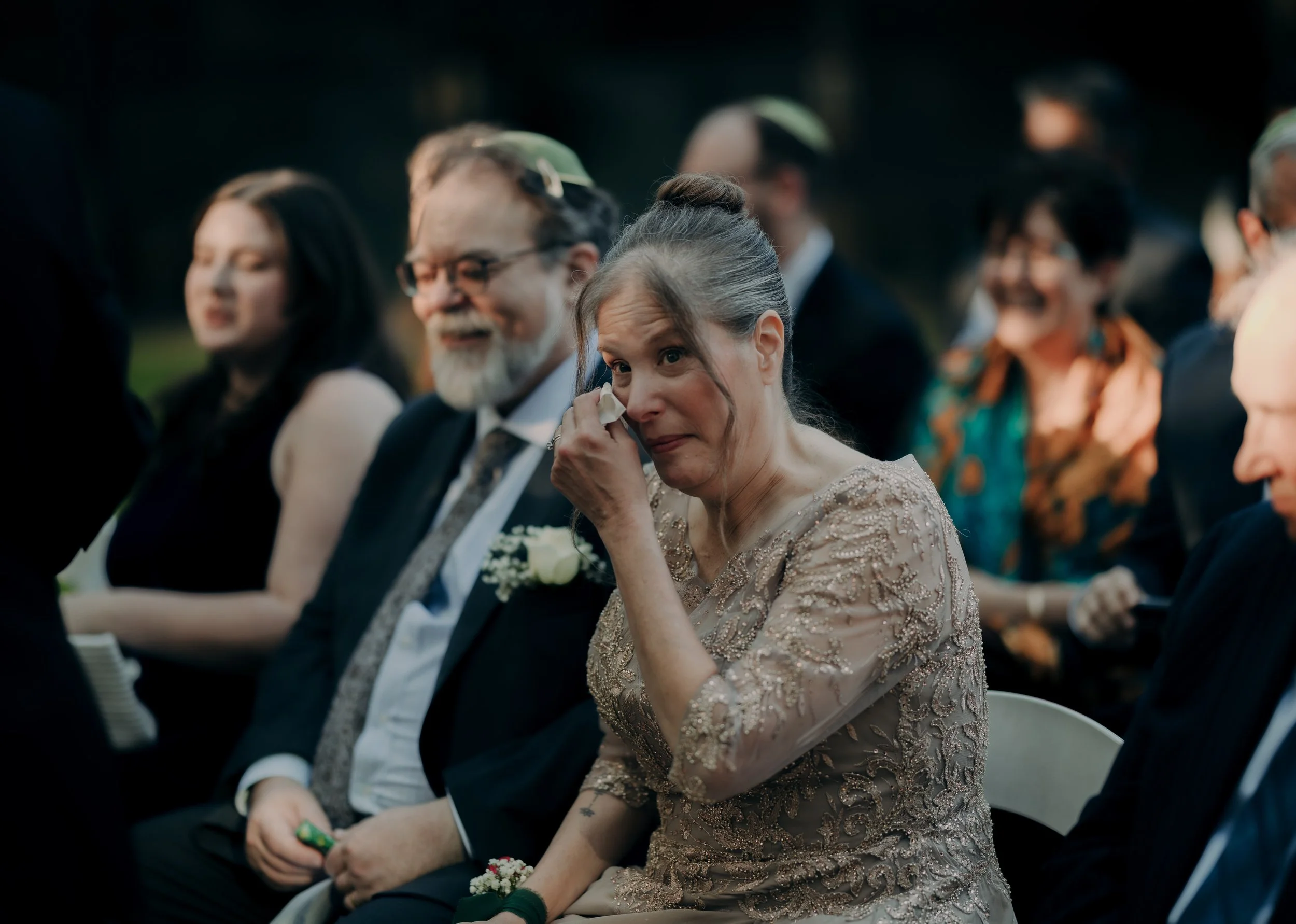 An older woman in a light pink gown, wiping tears from her eyes at an outdoor wedding ceremony, surrounded by seated guests.