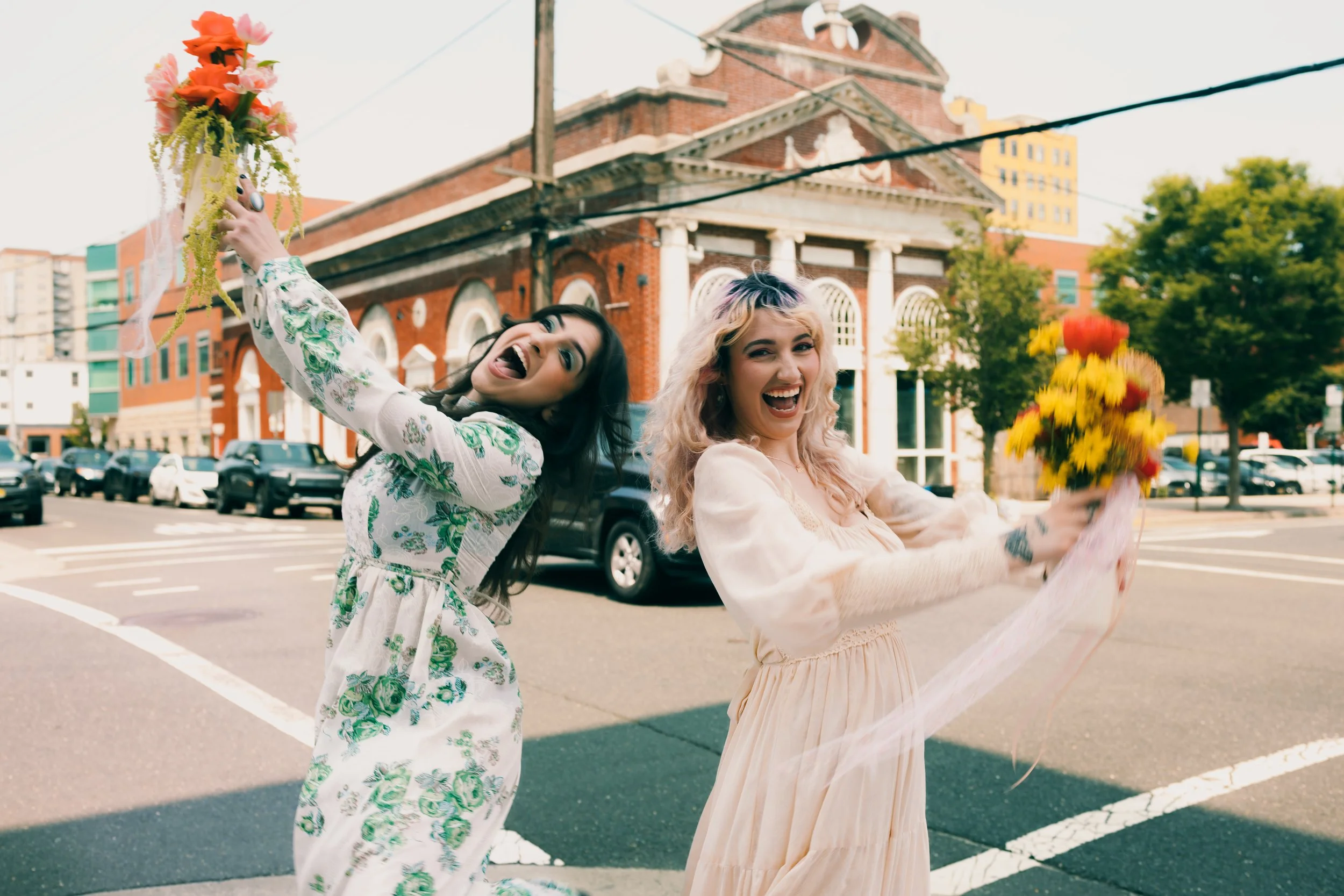 Two women smiling and holding bouquets of flowers, standing on a city street with historic brick buildings and parked cars in the background.