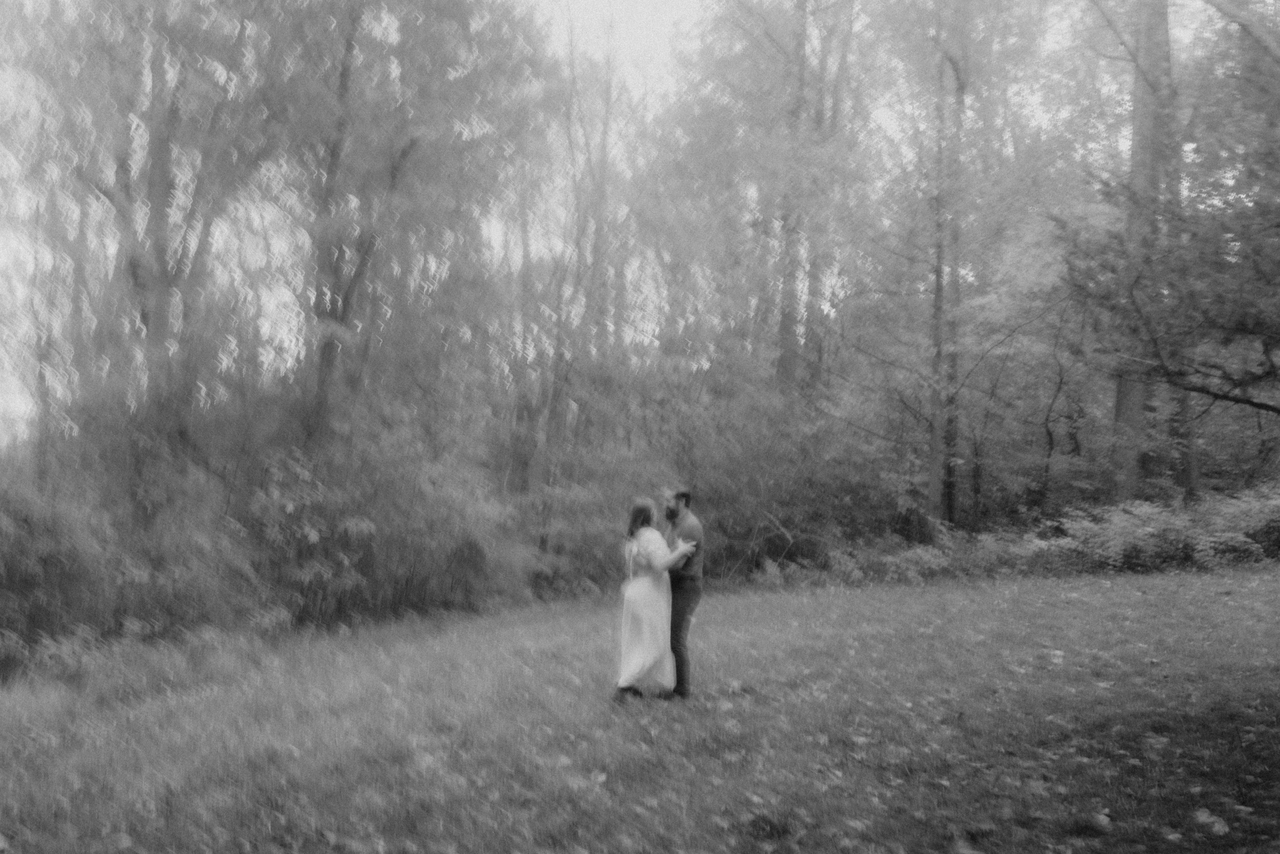 A black and white photo of a couple standing closely together in a natural outdoor setting with trees and grass, possibly in a park or wooded area.