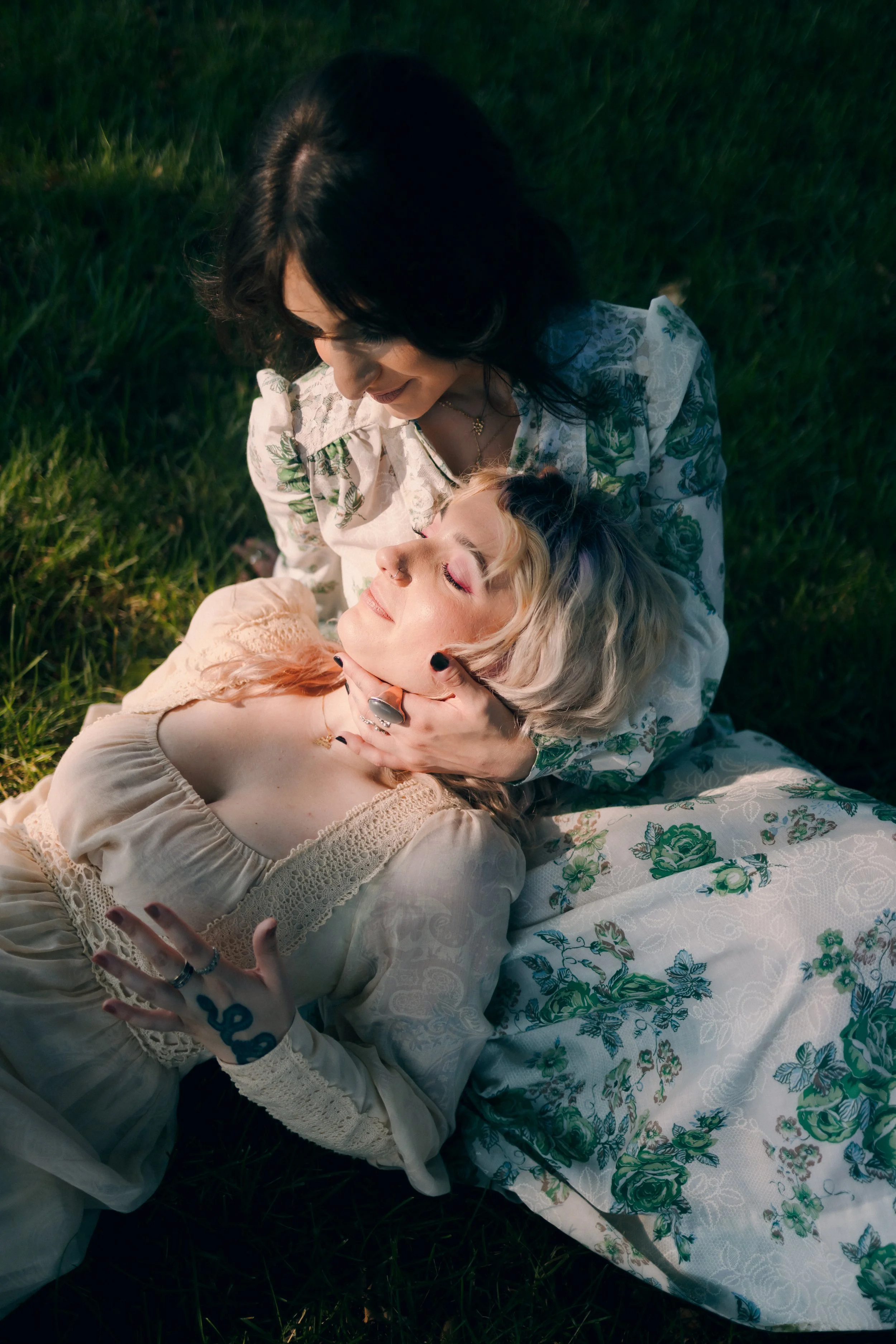 Two women, one with dark hair and the other with blonde hair, sitting outdoors on grass, sharing an intimate moment. The dark-haired woman gently holds the face of the blonde woman while they look at each other affectionately, surrounded by natural l