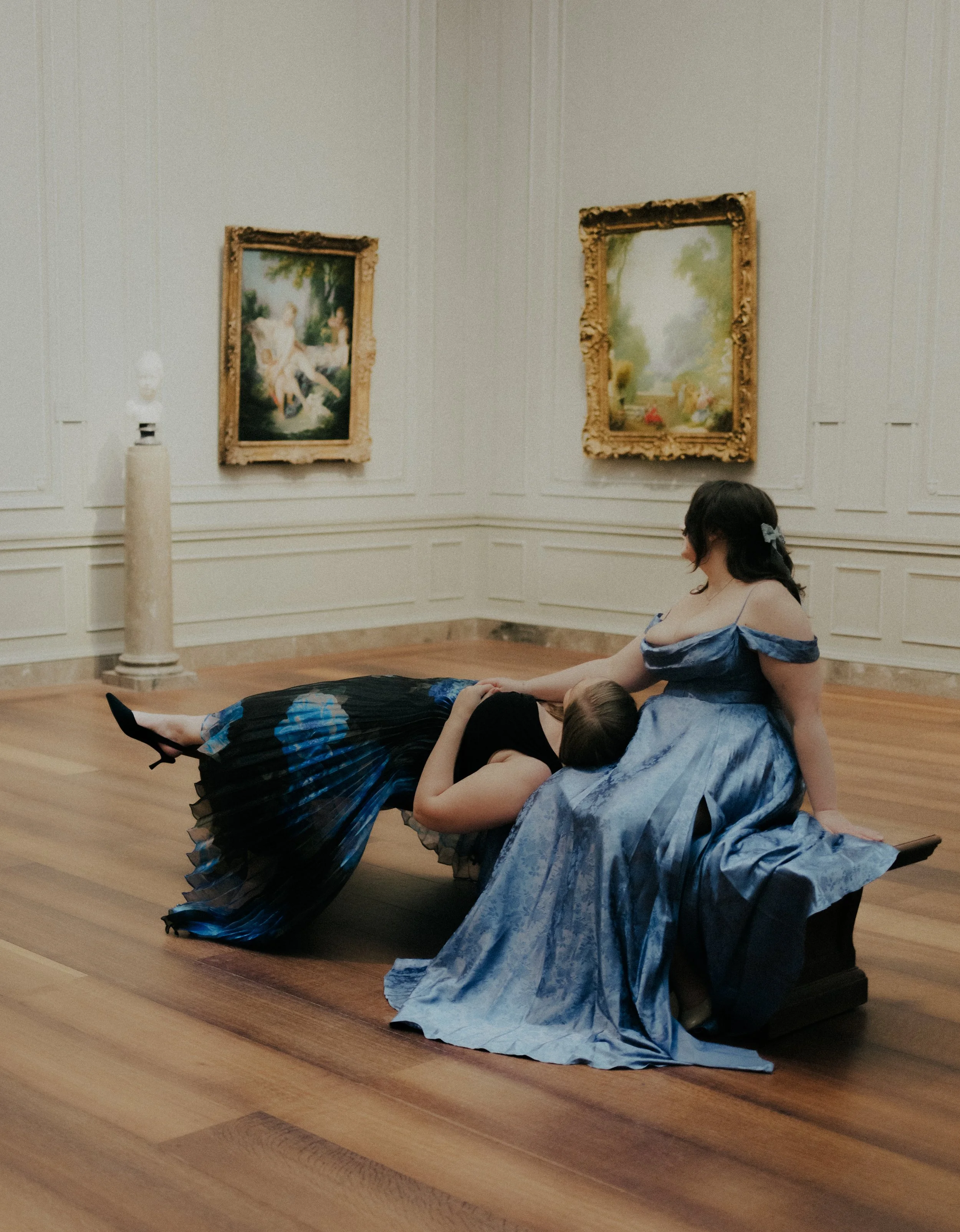 Two women in elegant dresses sitting and lying on a bench in an art gallery, with framed paintings on the wall behind them.