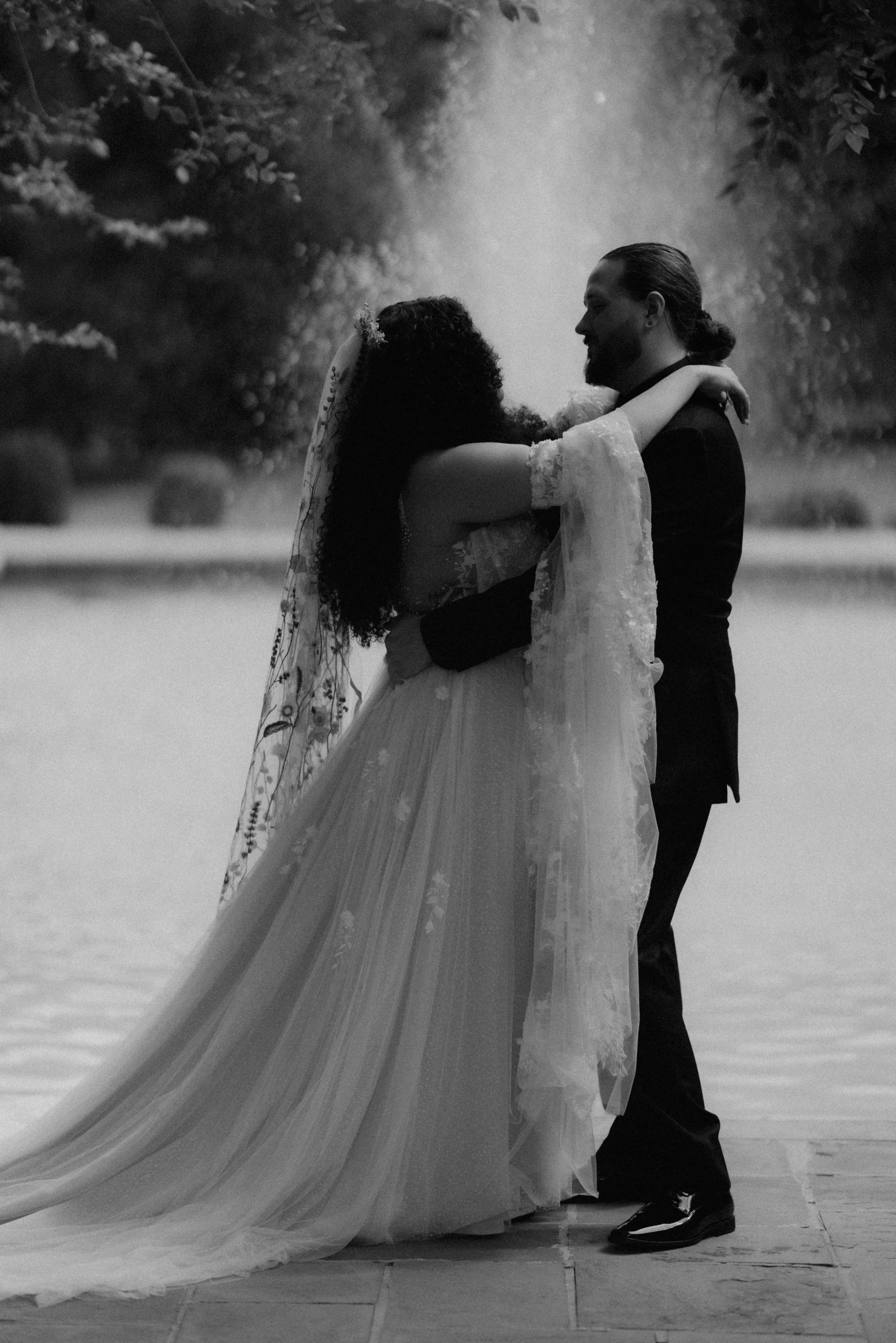 A bride and groom embrace outdoors in black and white, with the bride in a flowing wedding dress and veil, and the groom in a suit, near a waterfall or misty background.