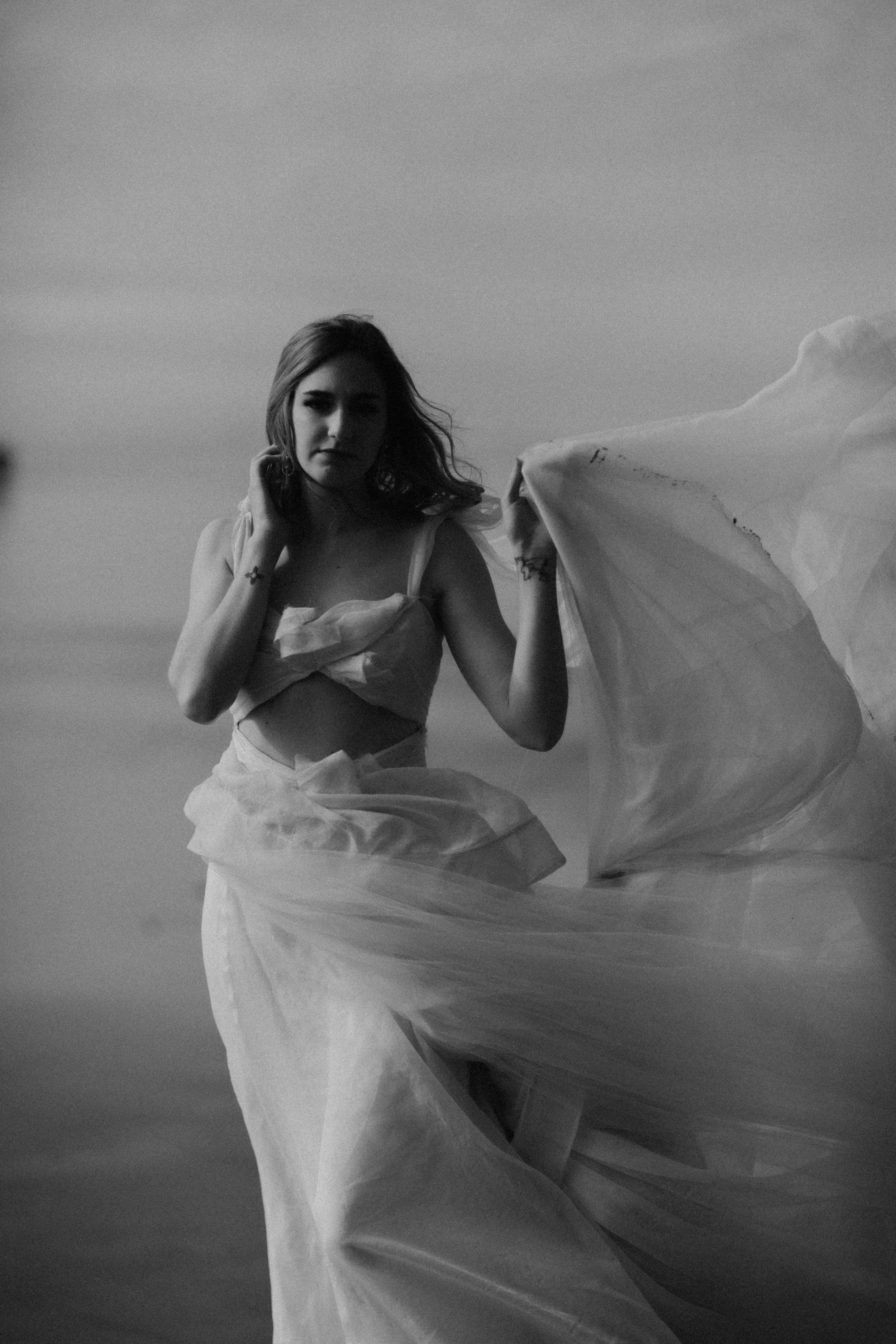 A black and white photo of a woman wearing a flowing skirt and a top, standing outdoors, with her hand touching her hair and looking at the camera.