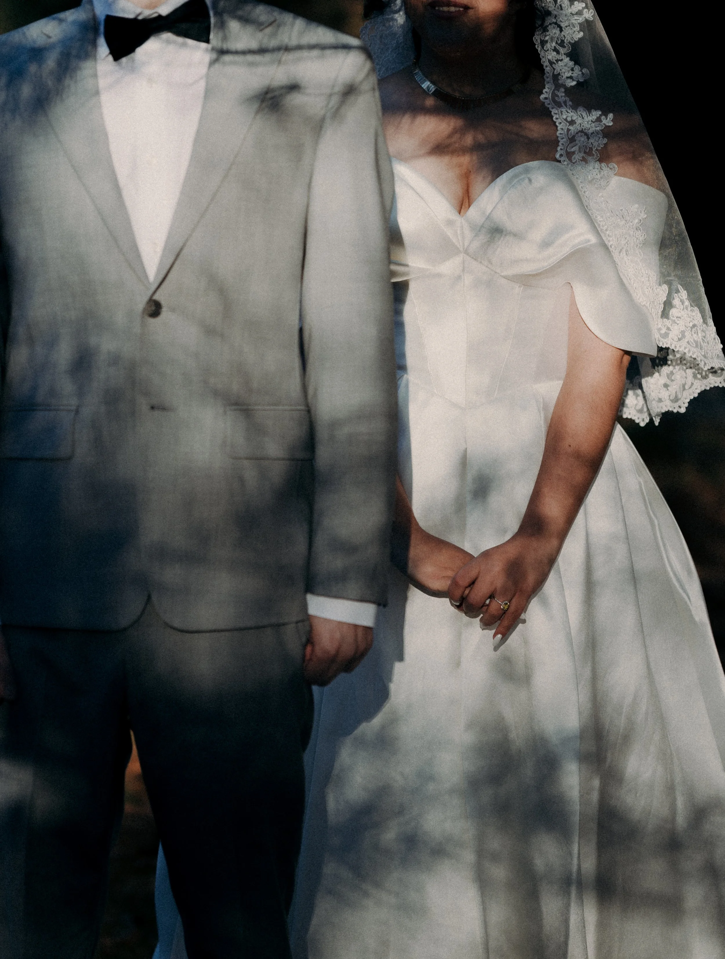 Close-up of a bride and groom holding hands, with the groom wearing a gray suit and bow tie, and the bride in a white wedding dress with lace details and a veil.