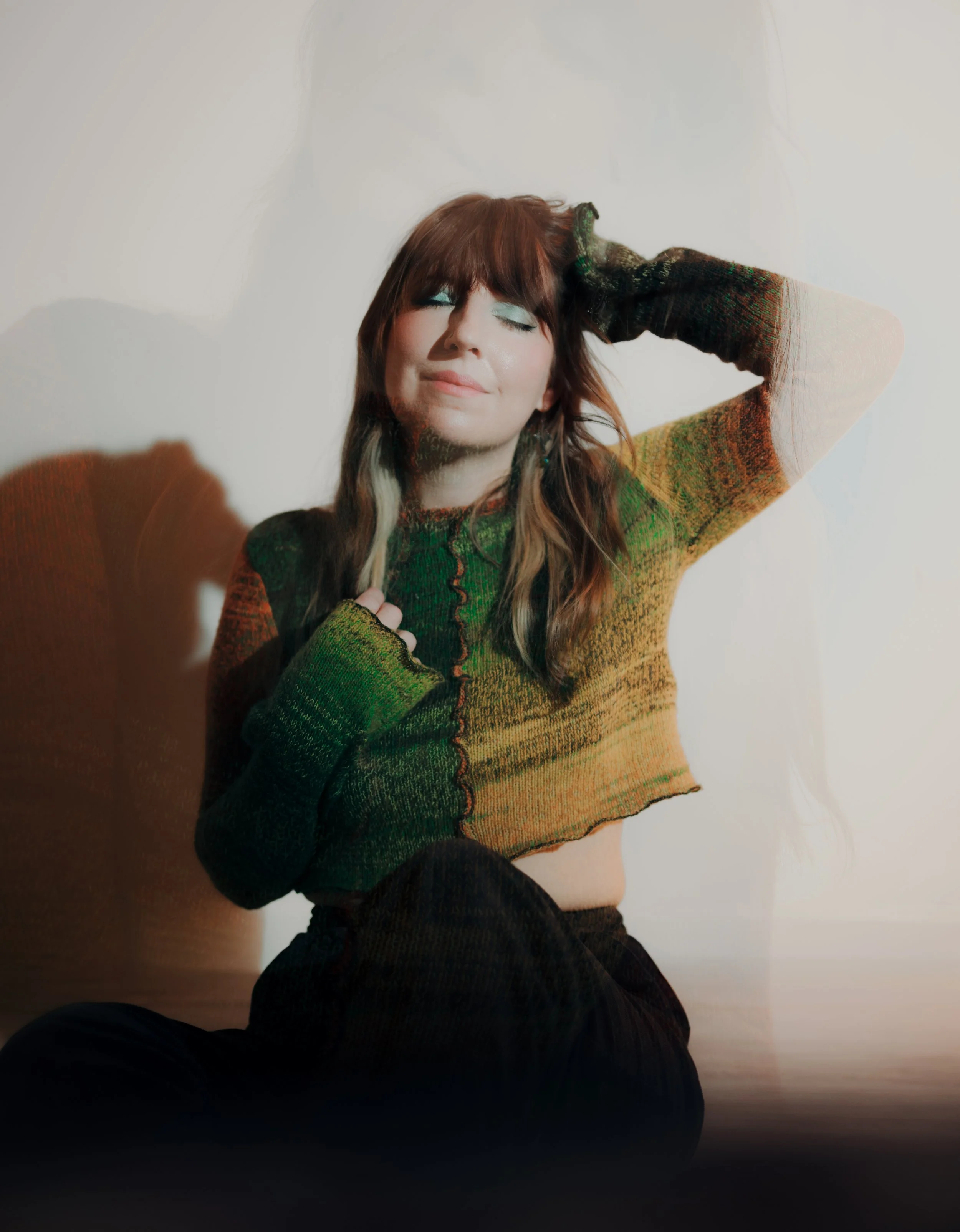 A woman with long brown hair, closed eyes, and a peaceful expression, wearing a multicolored knit sweater, sitting against a plain white background with a slight shadow, and her right hand resting on her head.