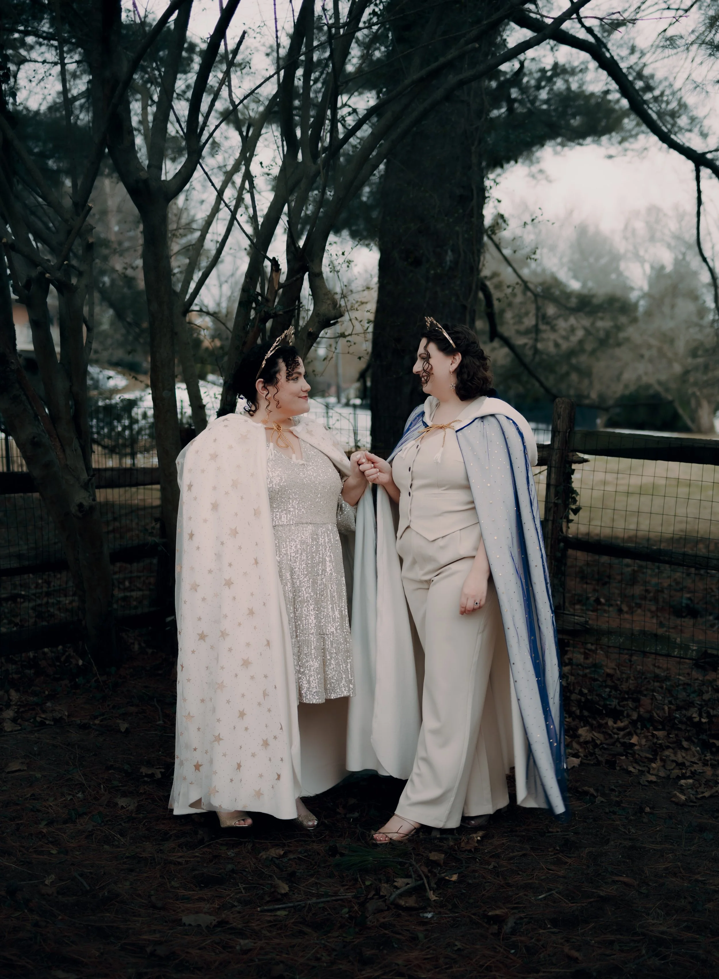 Two women dressed as fairies, wearing capes with star and stripe patterns, standing outdoors under leafless trees, holding hands and smiling at each other.