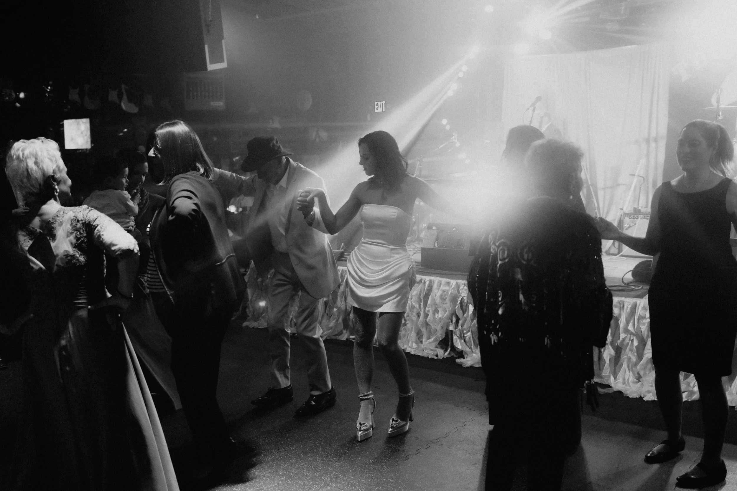 People dancing and enjoying music at a party or nightclub, with a DJ booth and stage in the background.