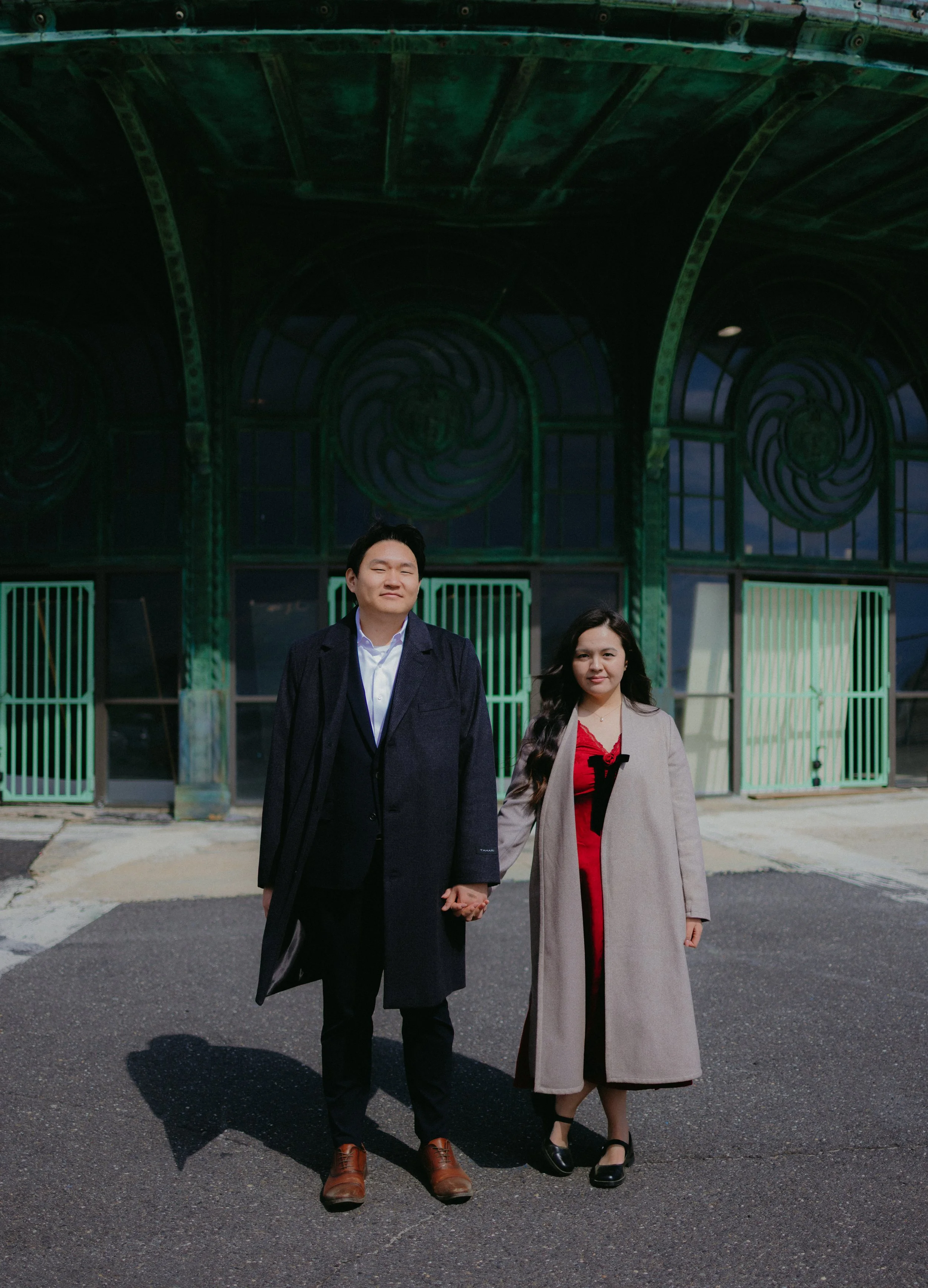 A couple holding hands on a sunny day, looking directly into the camera, in front of the Asbury Park carousel.
