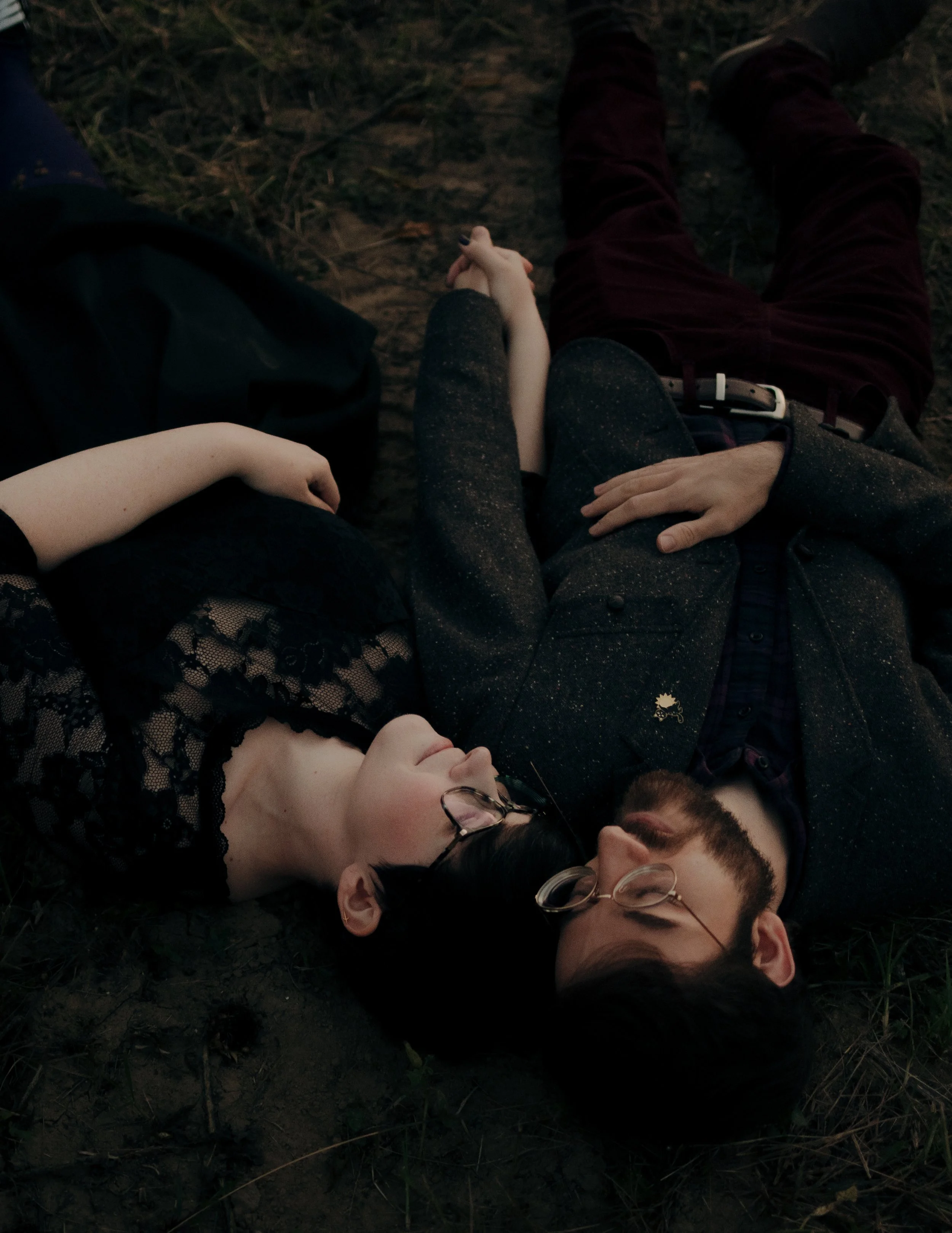 A woman and a man are lying on the ground outdoors, holding hands and looking at each other. The woman is wearing a black dress with lace, and the man is wearing a dark speckled blazer, glasses, and has a beard.