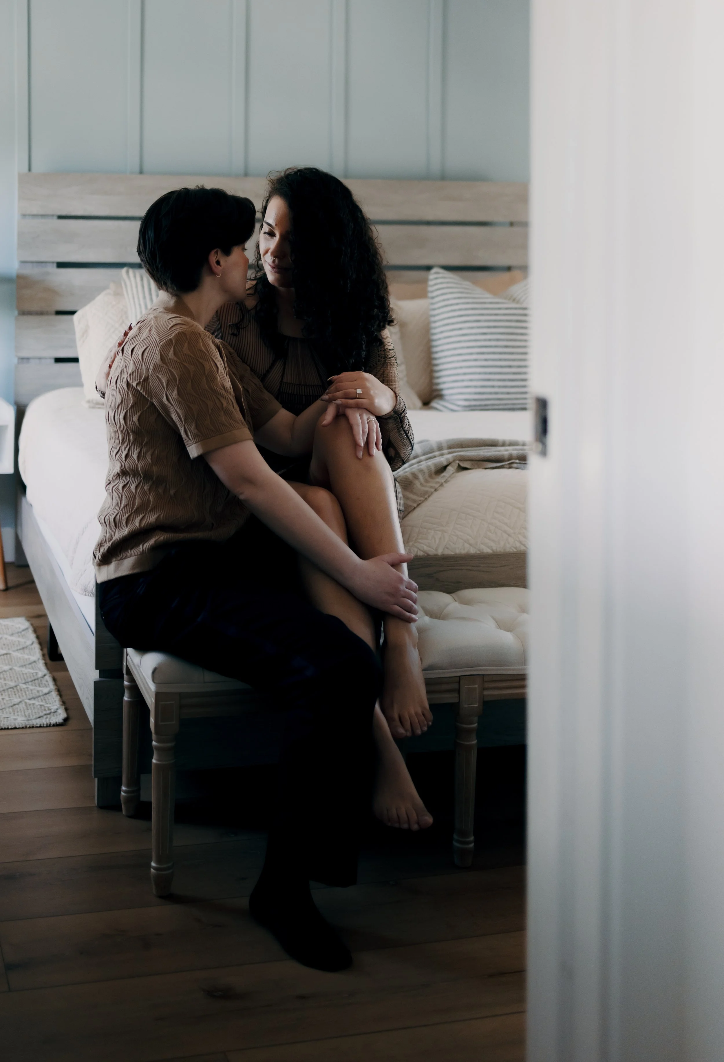 Two women sit on a bench at the foot of a bed, engaging in an intimate moment.