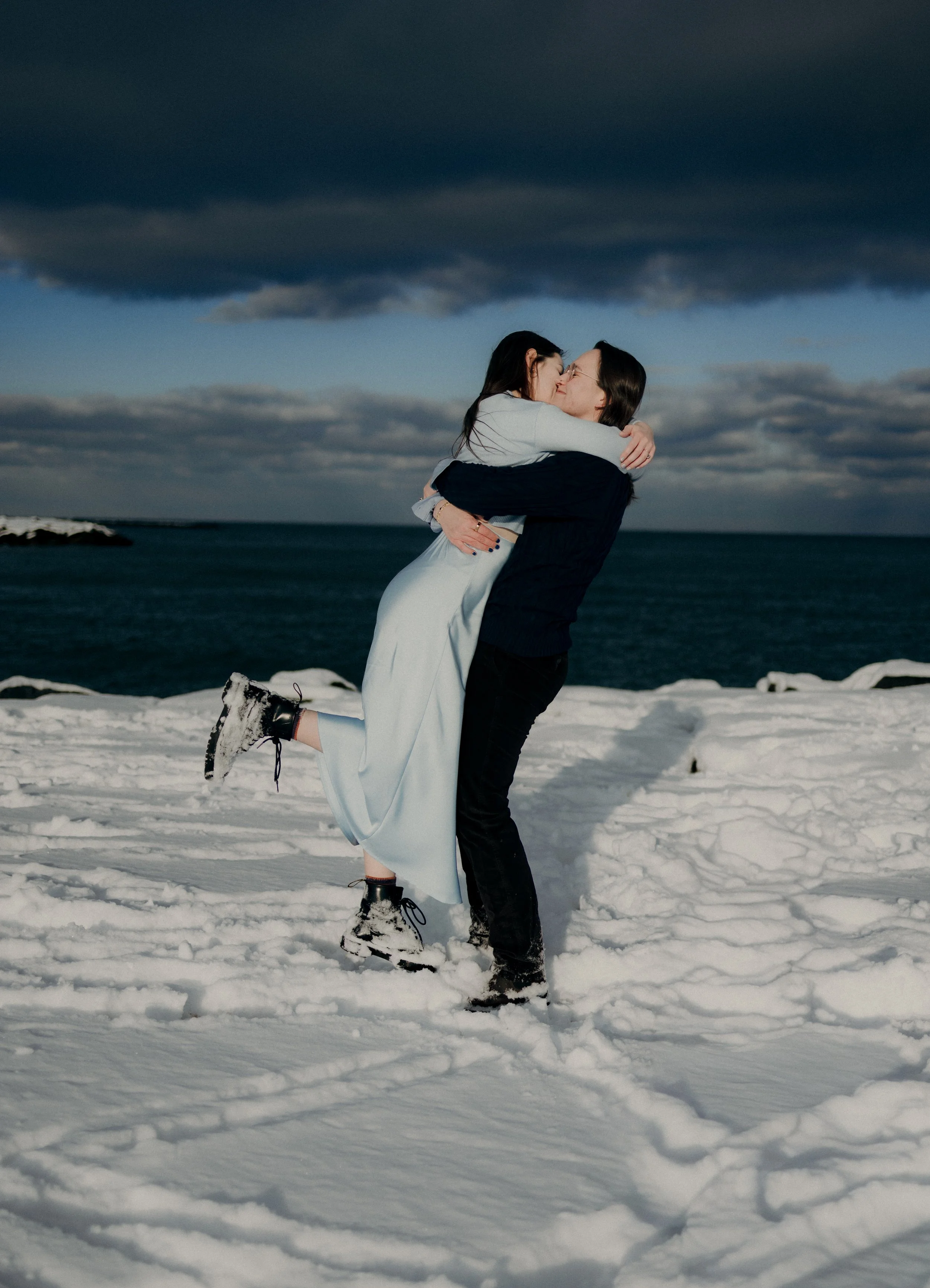 A couple sharing a kiss in the snow with a dark cloudy sky and ocean in the background.