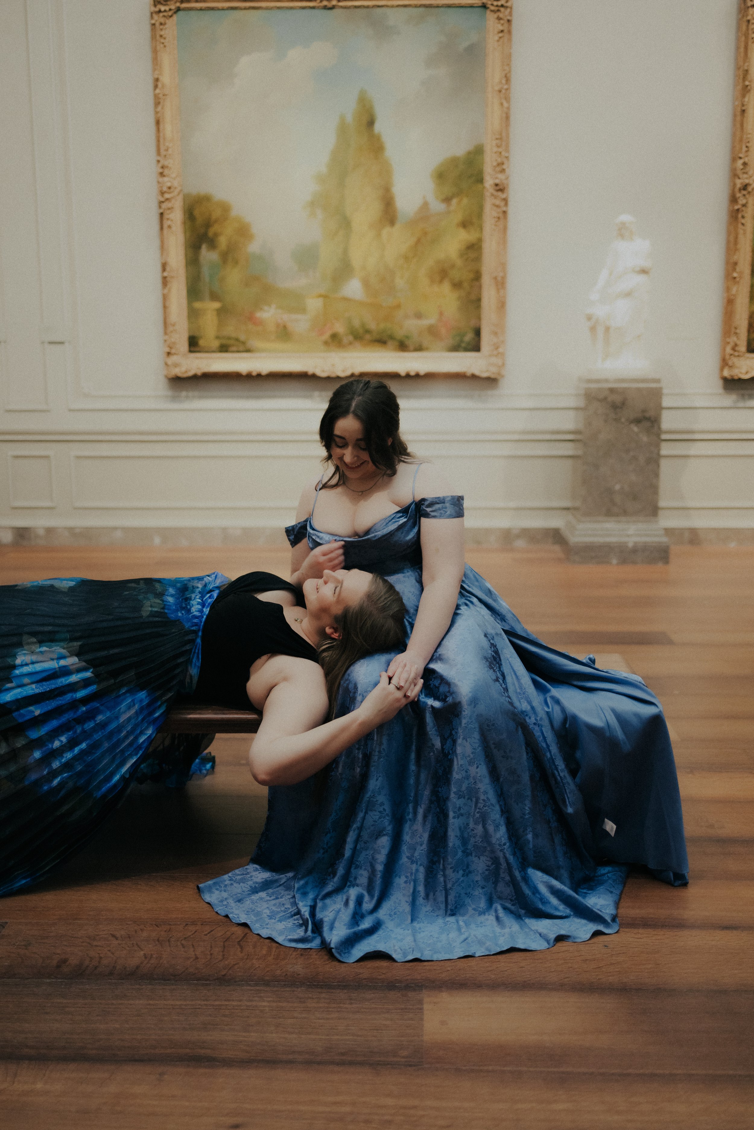 Two women wearing evening gowns, one seated and the other lying down on her lap, sharing a moment of joy in an art gallery with paintings and sculptures in the background.