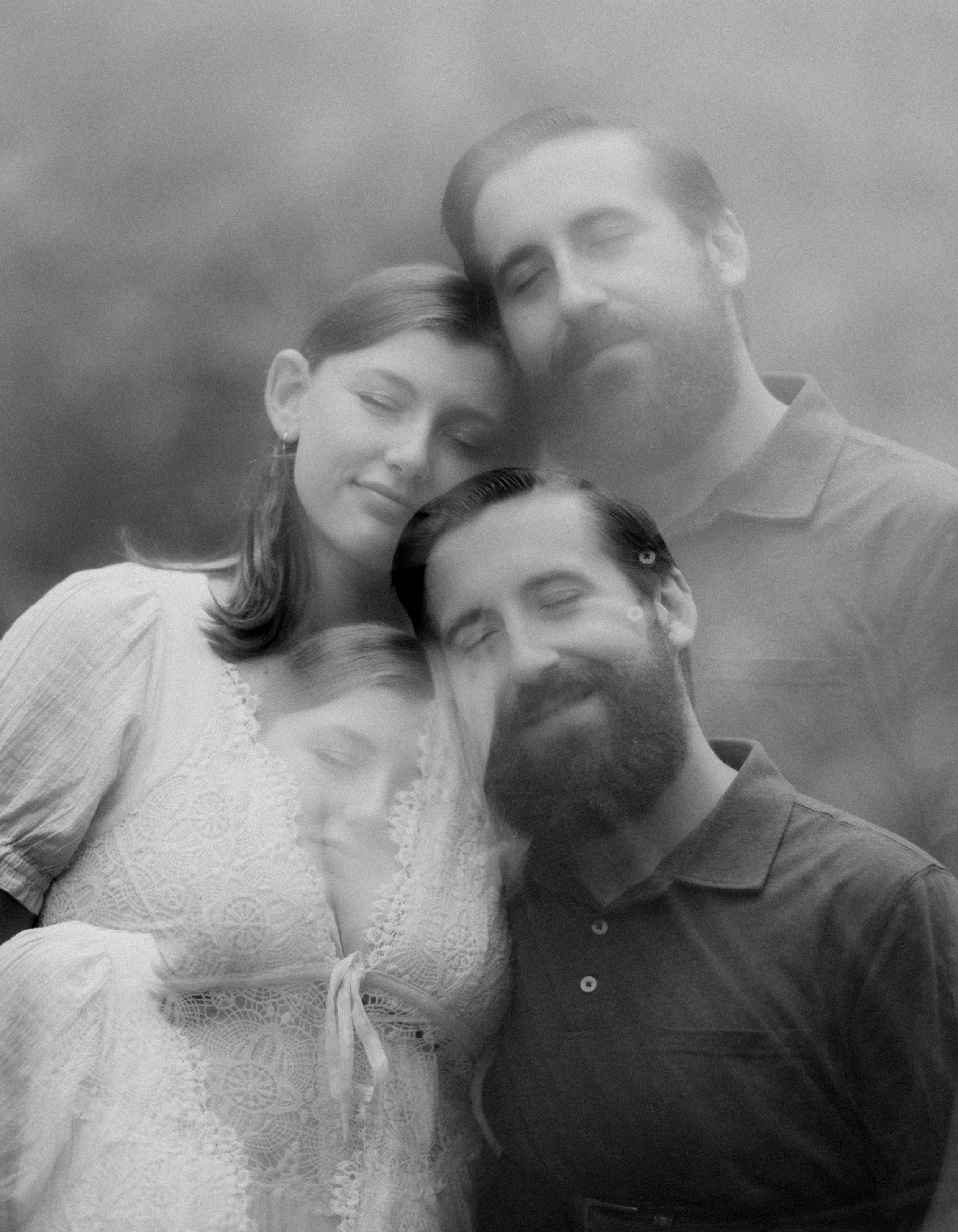 Black and white photo of four people with closed eyes, two men and two women, close together with heads touching, a foggy or cloudy background.