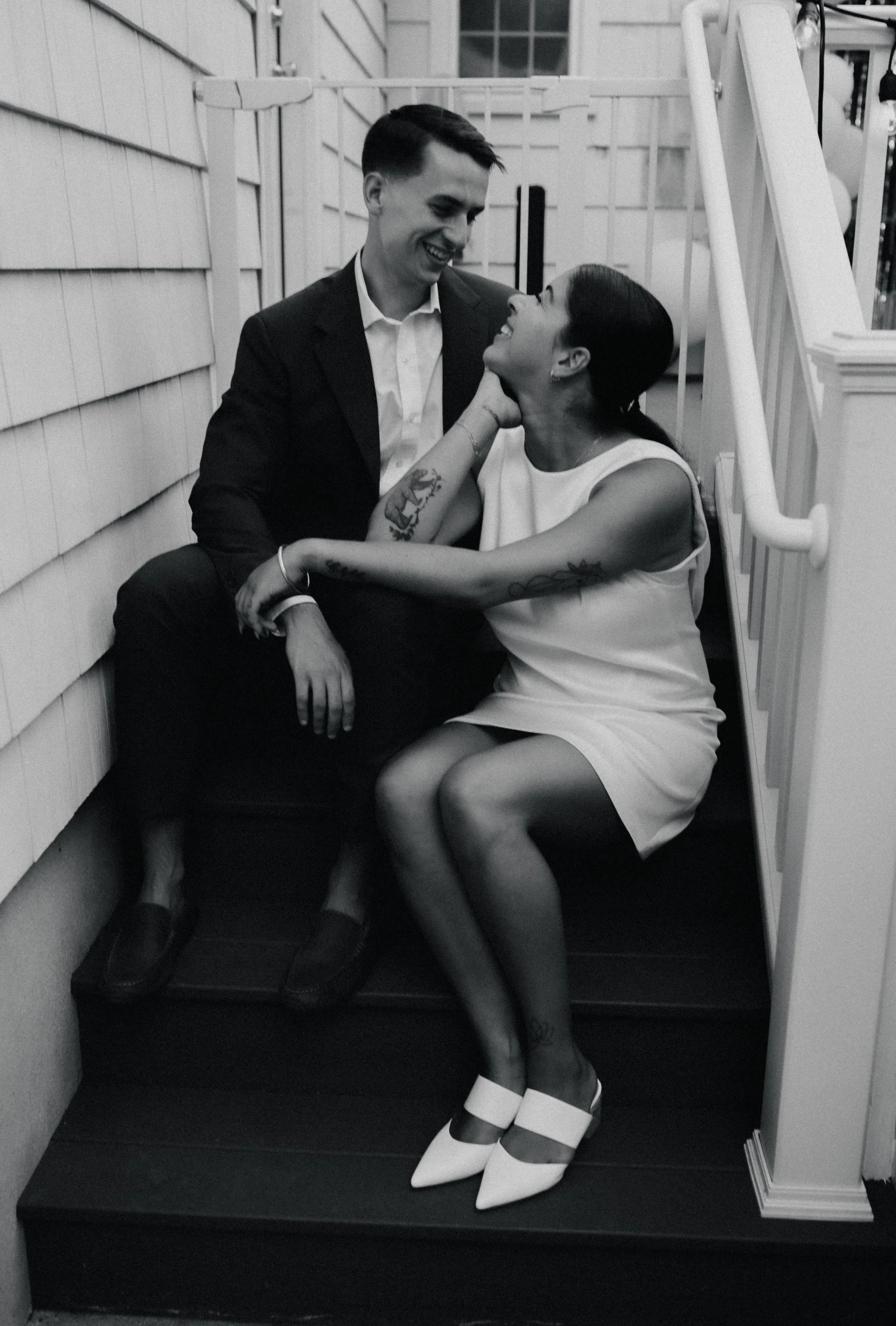 A black and white photo of a couple sitting on stairs, smiling and looking into each other's eyes. The man is in a suit, and the woman is wearing a sleeveless dress and pointed shoes.
