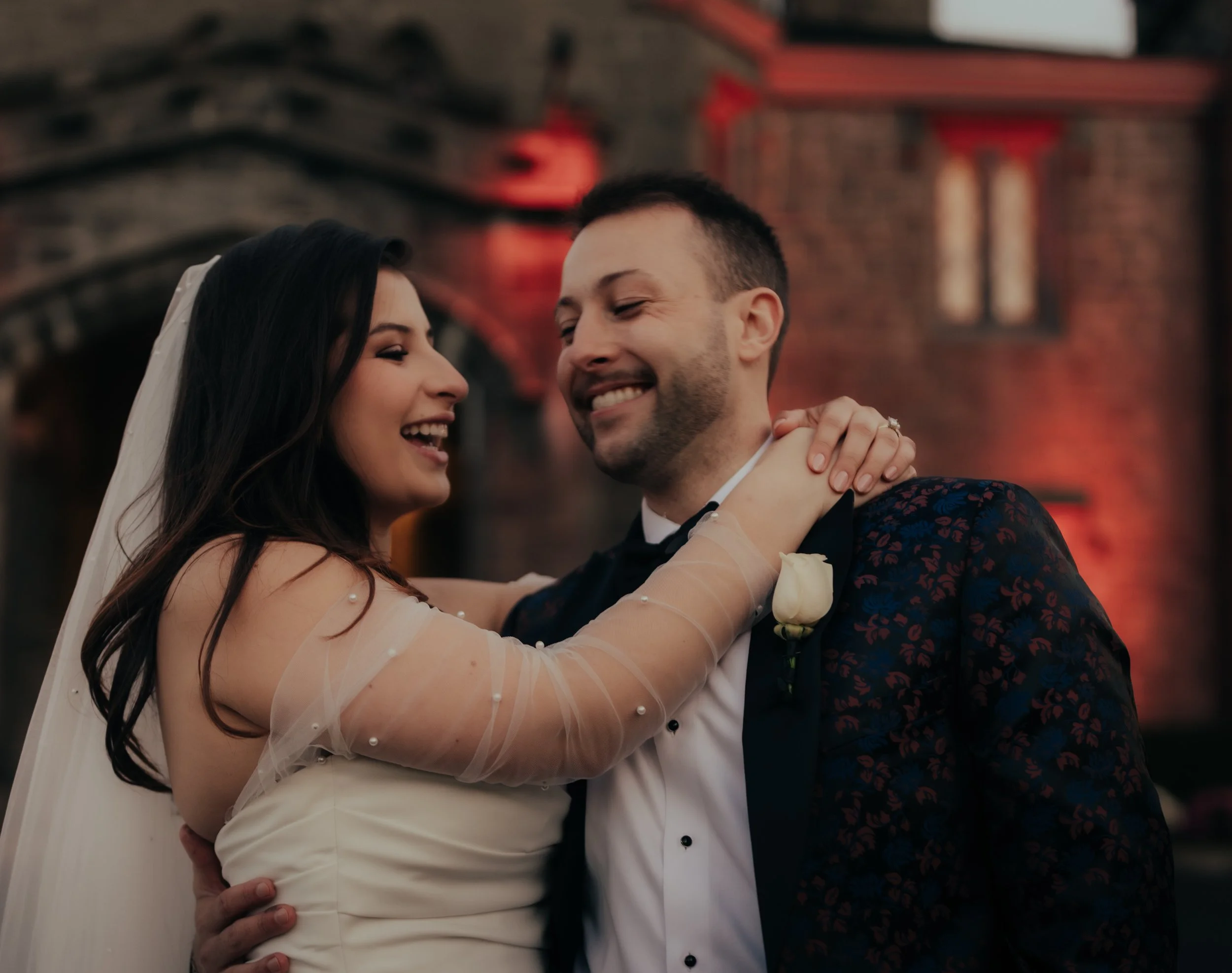 A bride and groom share a joyful dance outdoors at their wedding, with warm lighting illuminating a brick building in the background.