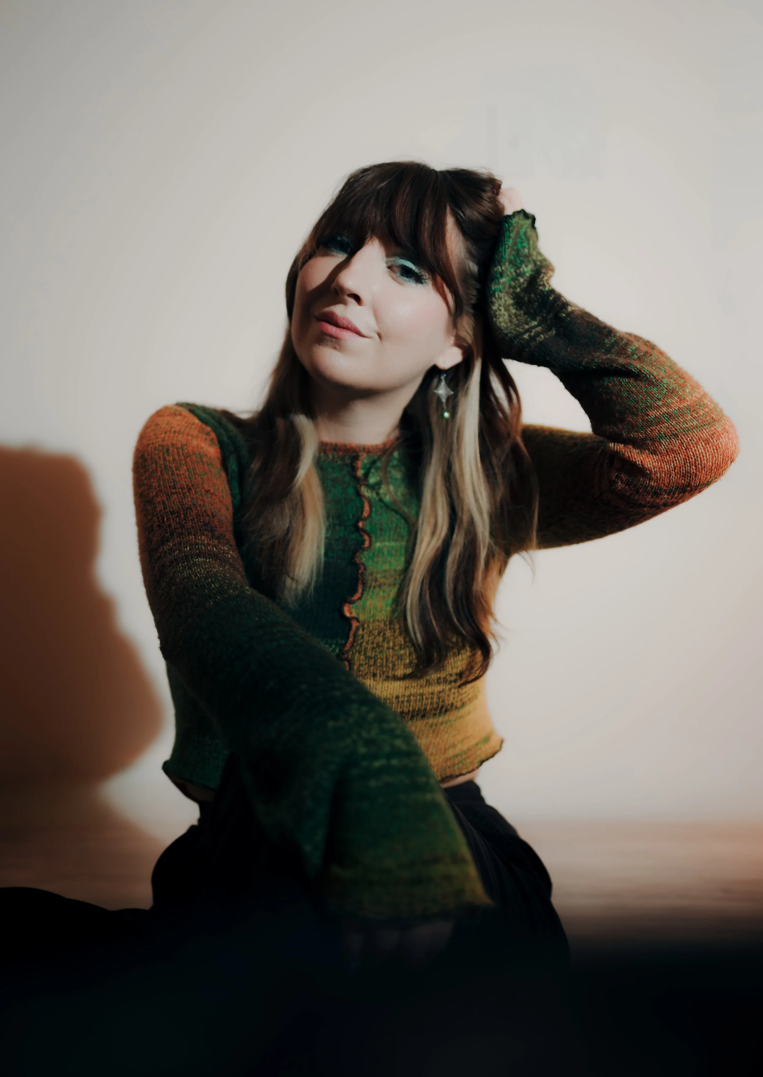 A woman with long brown hair and bangs, wearing a multicolored sweater with green, orange, and brown shades, sitting on the floor with one arm behind her head, smiling softly.