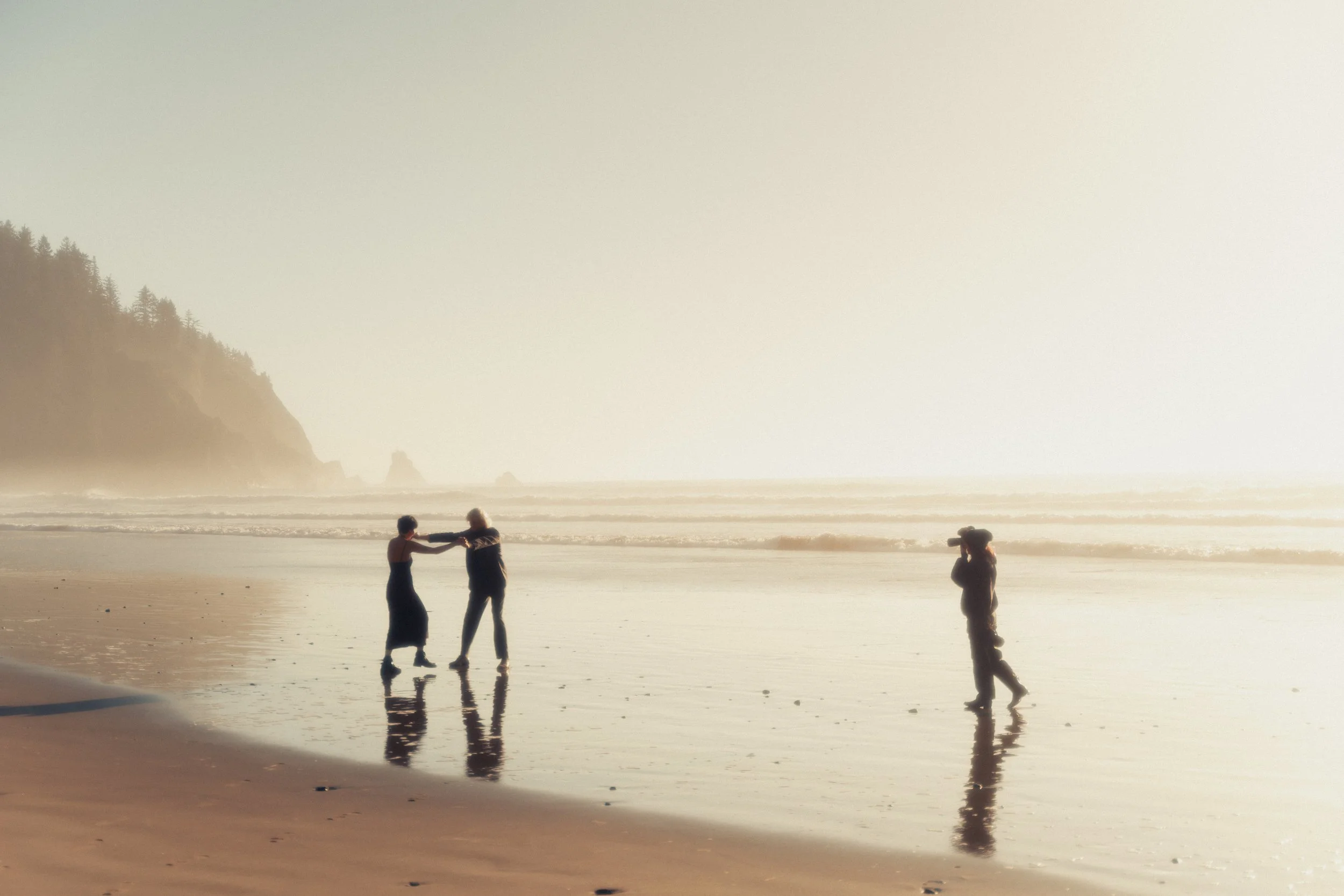 Two women dancing on a beach at sunset, with a man walking nearby and a photographer taking pictures.