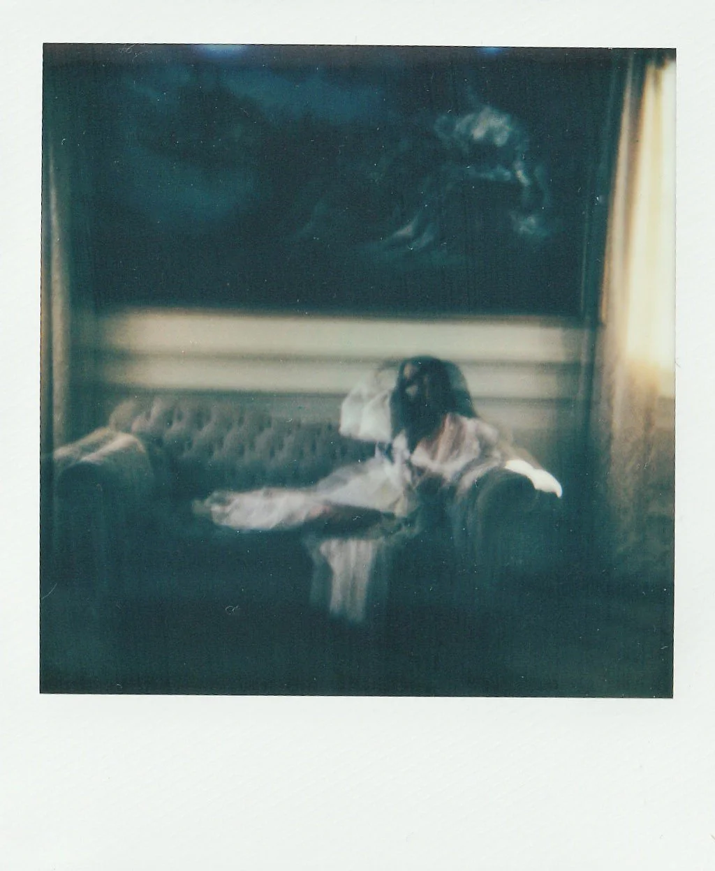 A woman dressed in vintage clothing seated on a couch in a dimly lit room, with a painting of a horse hanging above her.