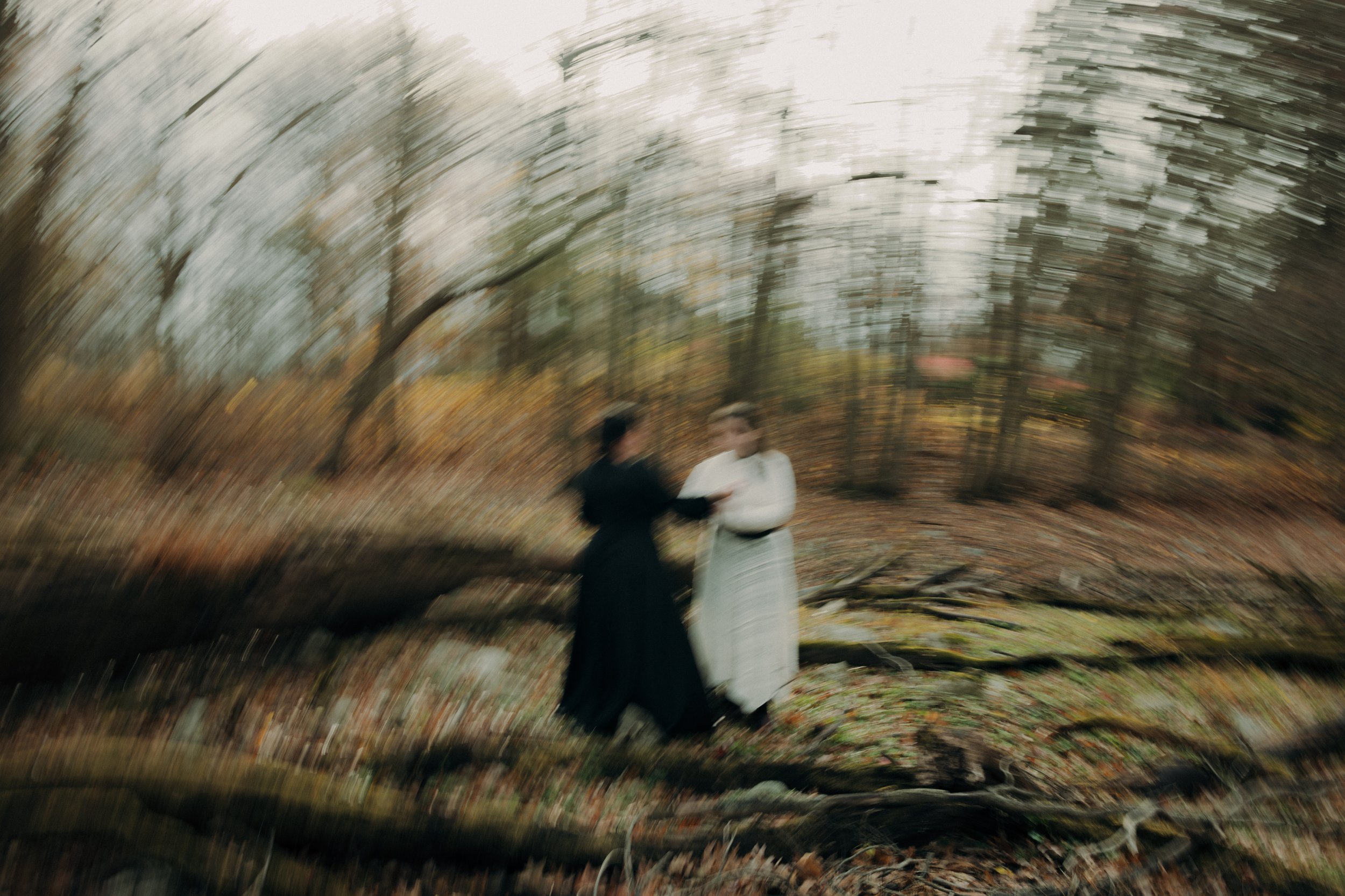 Two people dressed in vintage clothing standing in a forest with blurred trees and fallen leaves around them.