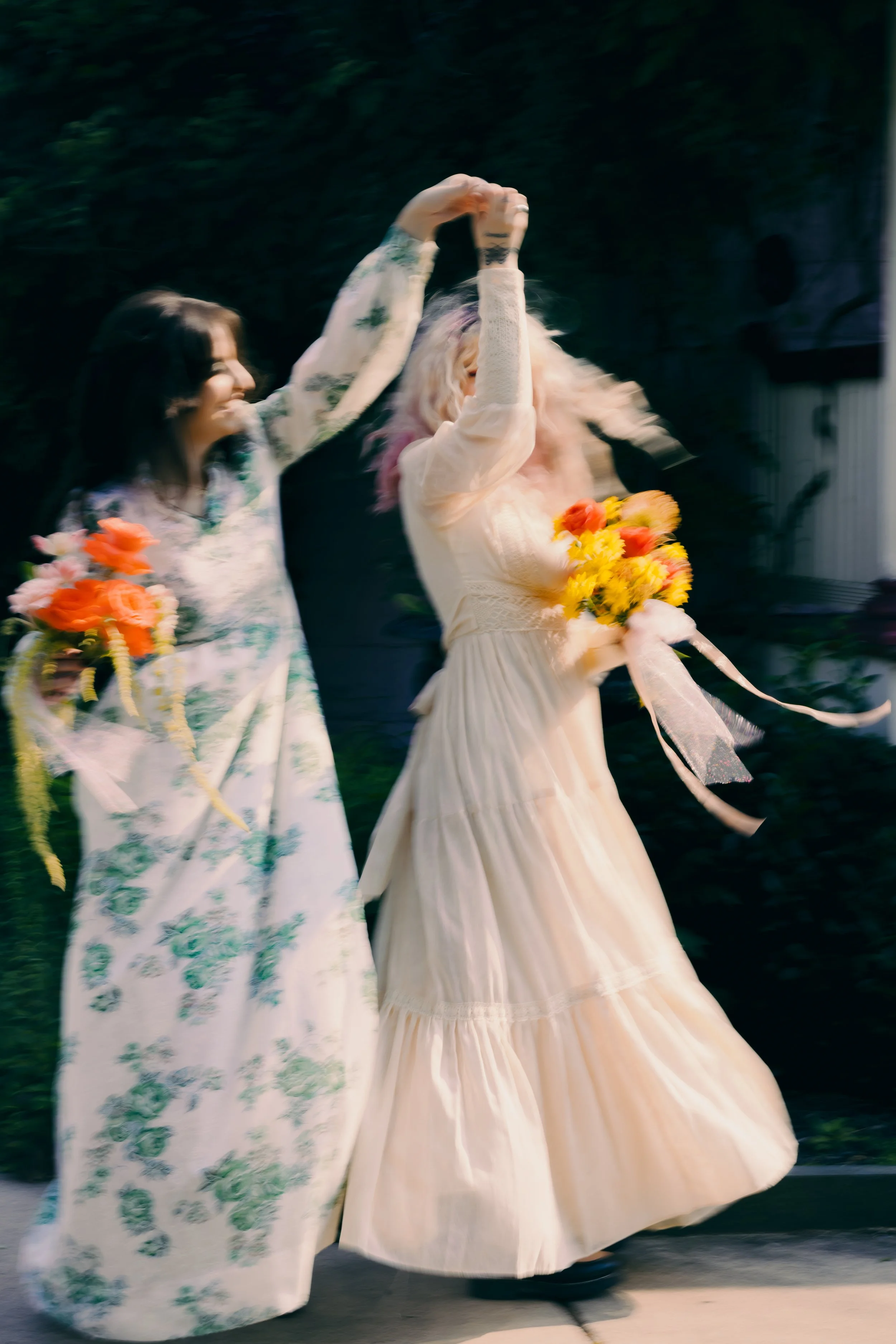 Two women wearing long dresses holding bouquets of flowers, dancing outdoors with blurred background.