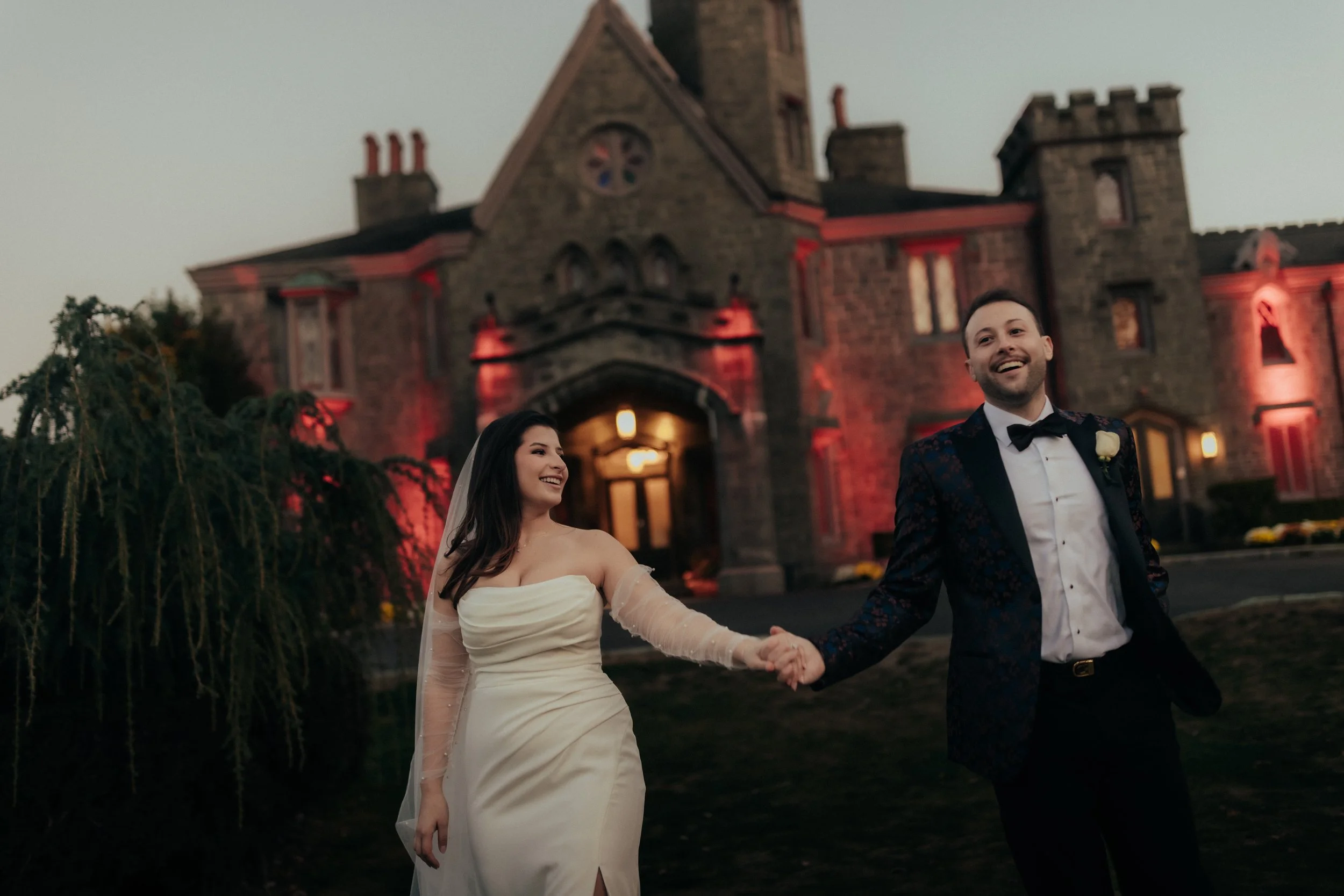 Bride and groom holding hands outside a mansion at dusk, with red lighting on the mansion.