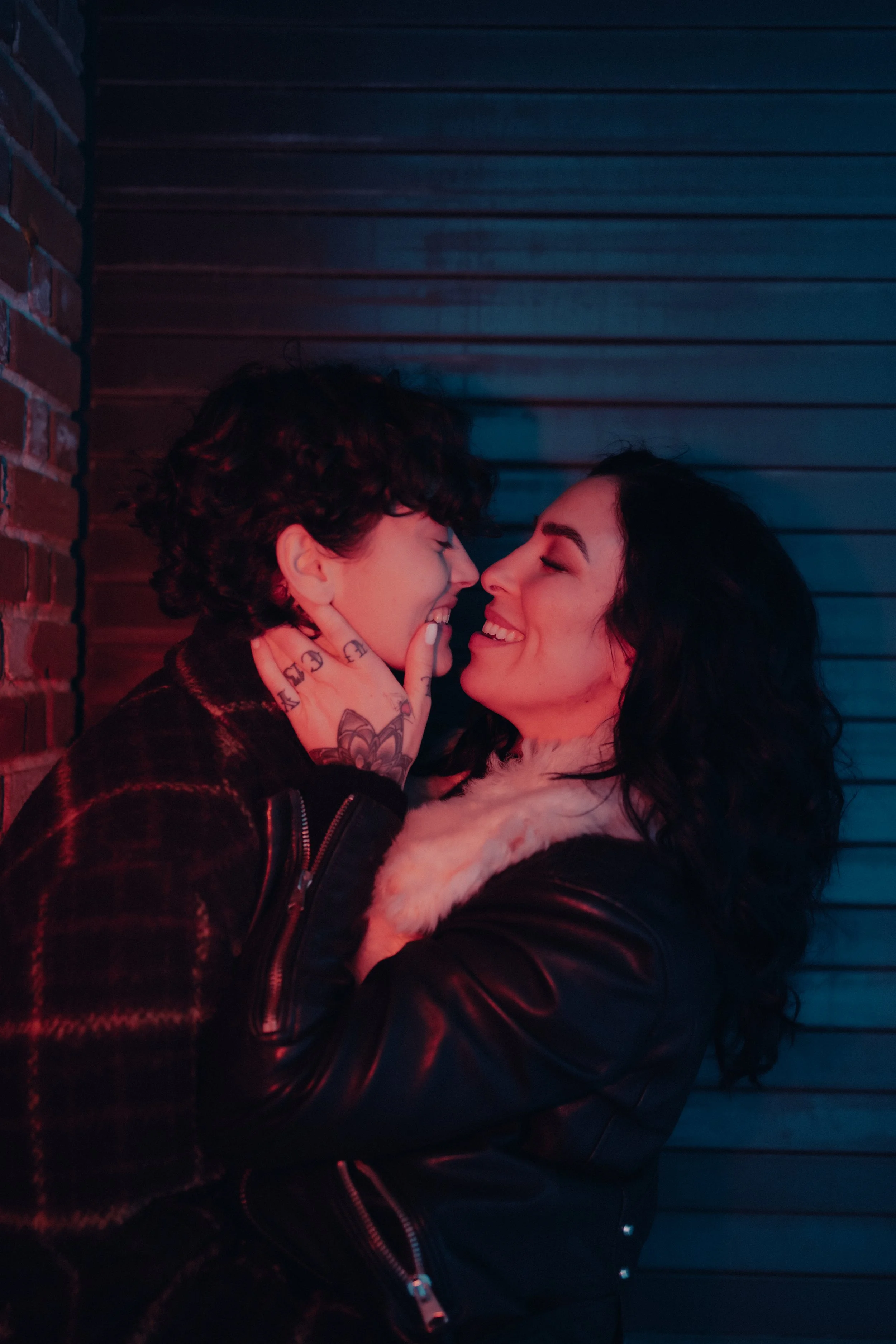 A couple sharing an intimate moment, with faces close and smiling, against a colored in urban alleyway background.