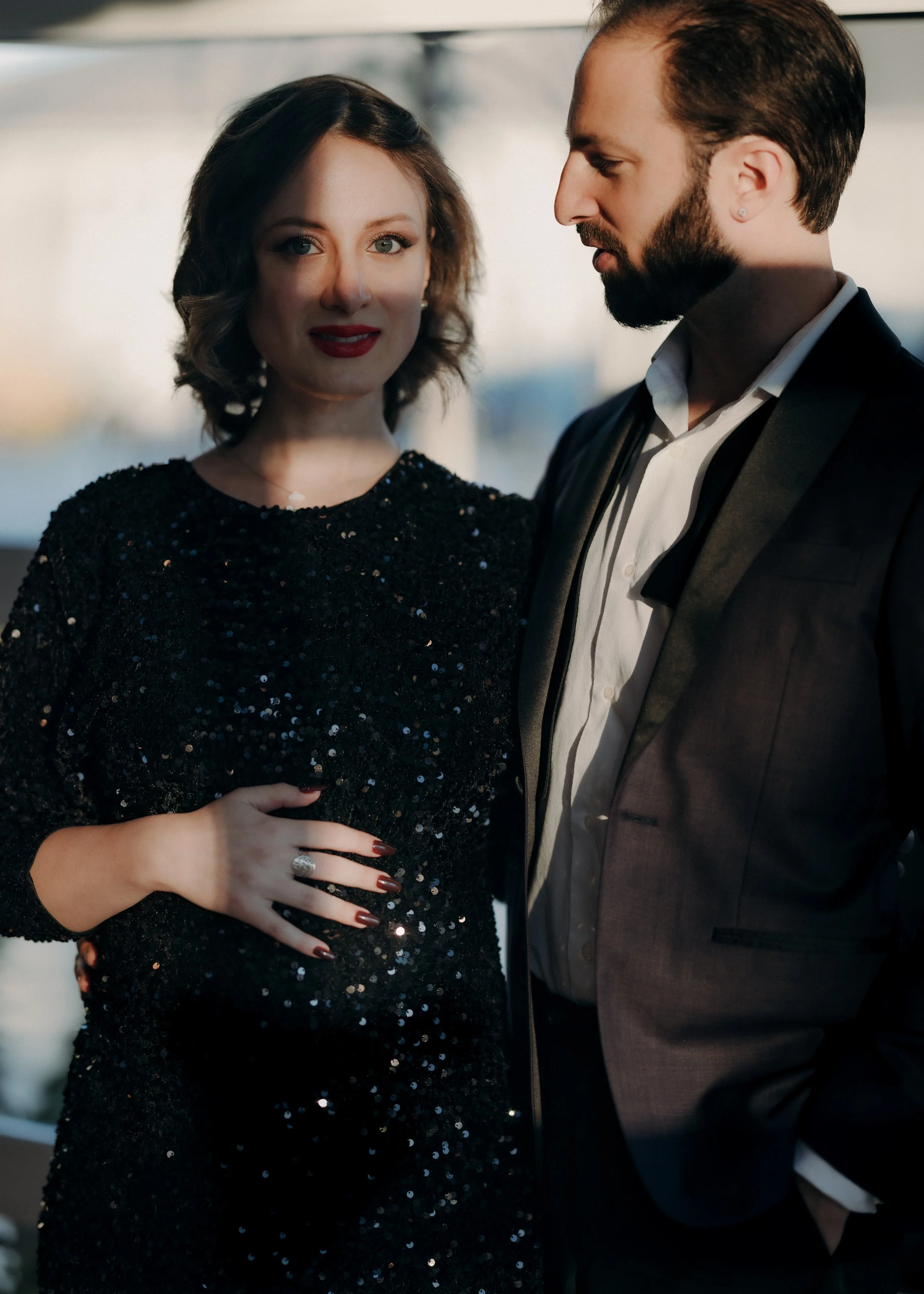 A woman wearing a black, sequined dress with red lipstick and dark nail polish, and a man with a beard in a suit, are standing close together indoors with a blurred background, possibly at an evening event.