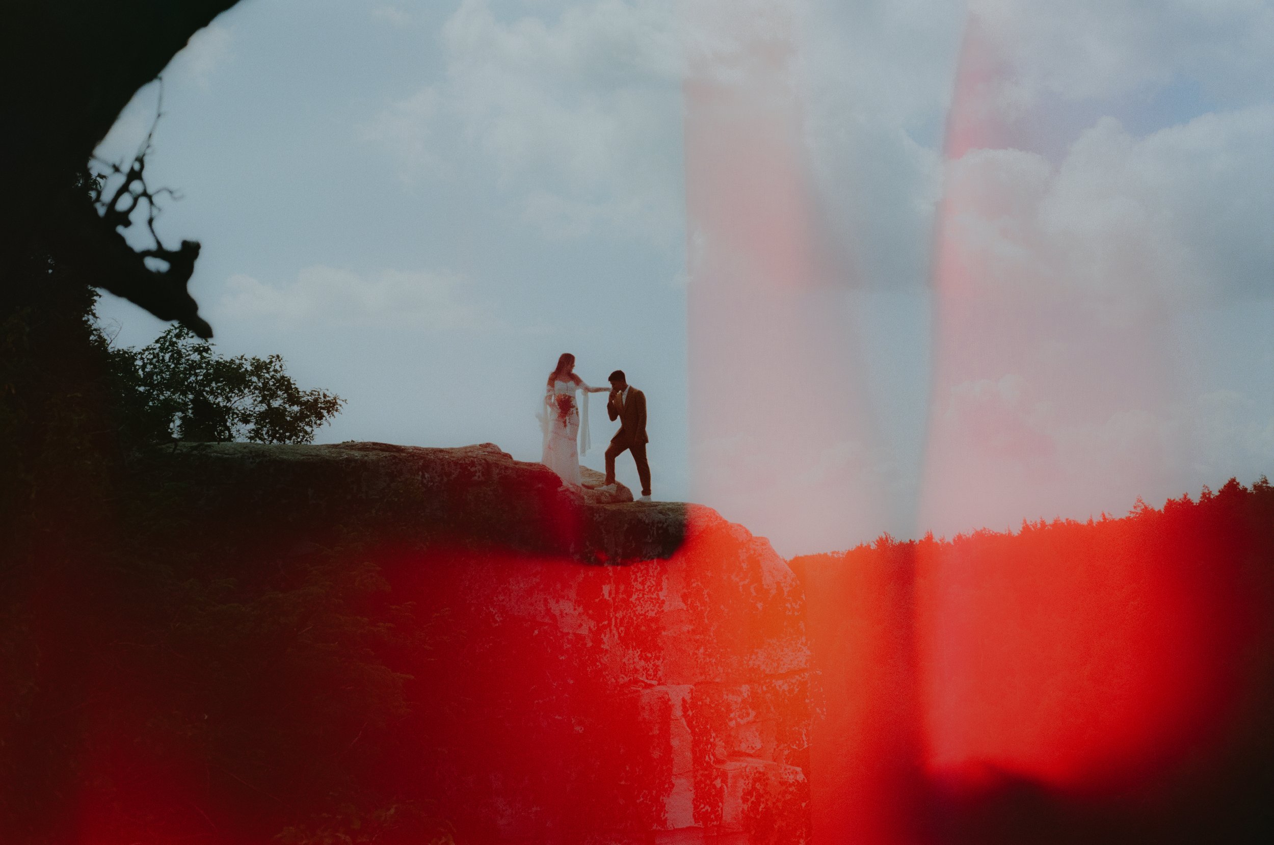 A couple stands on a rocky ledge, with the woman in a wedding dress and the man in a suit, holding hands against a cloudy sky background.
