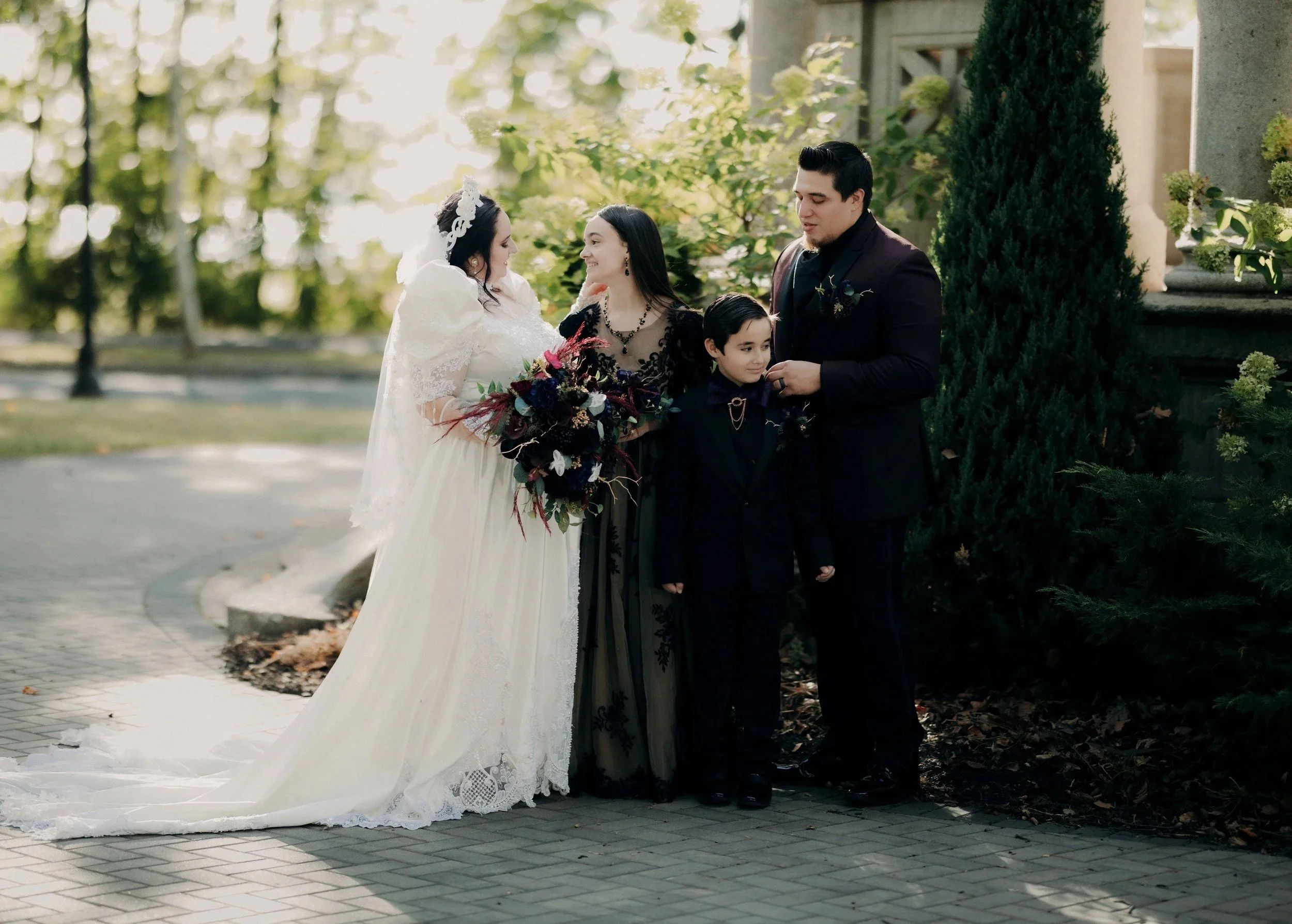 A wedding scene outdoors with a bride holding a bouquet of dark and colorful flowers, standing next to a woman in black dress, a young boy in a black suit, and a man in a black suit. They are smiling and talking, with greenery and trees in the backgr
