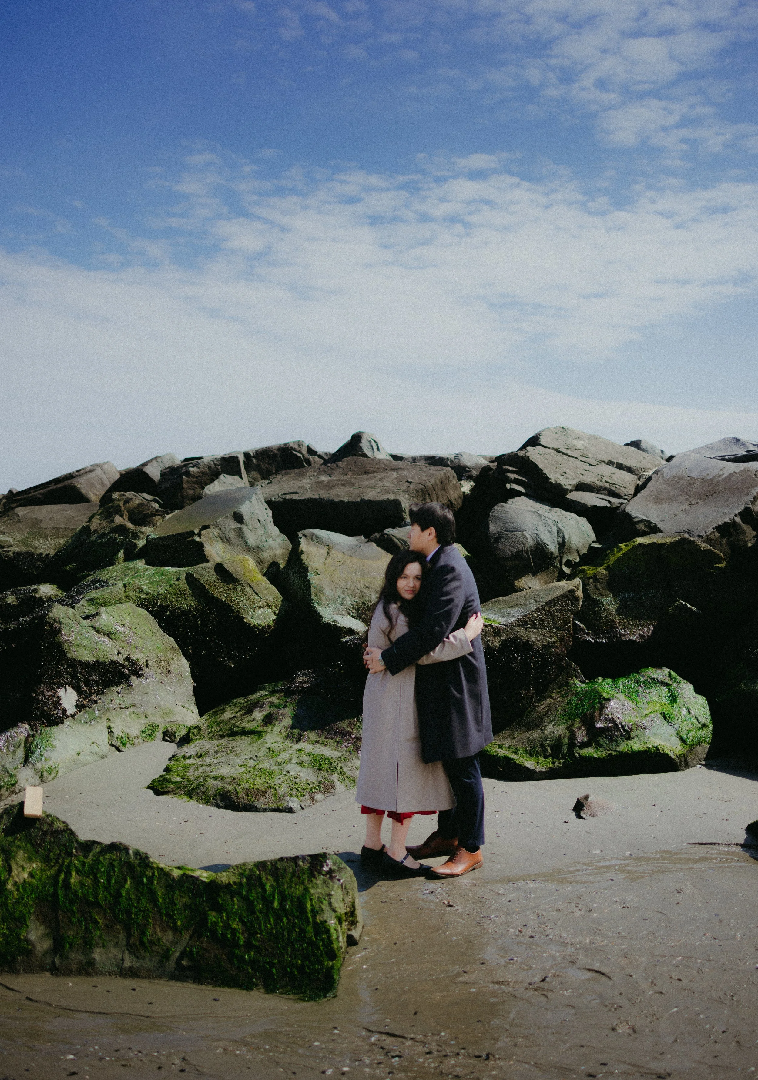 A couple hugging on a sandy beach with large rocks covered in green moss and a blue sky with scattered clouds in the background.