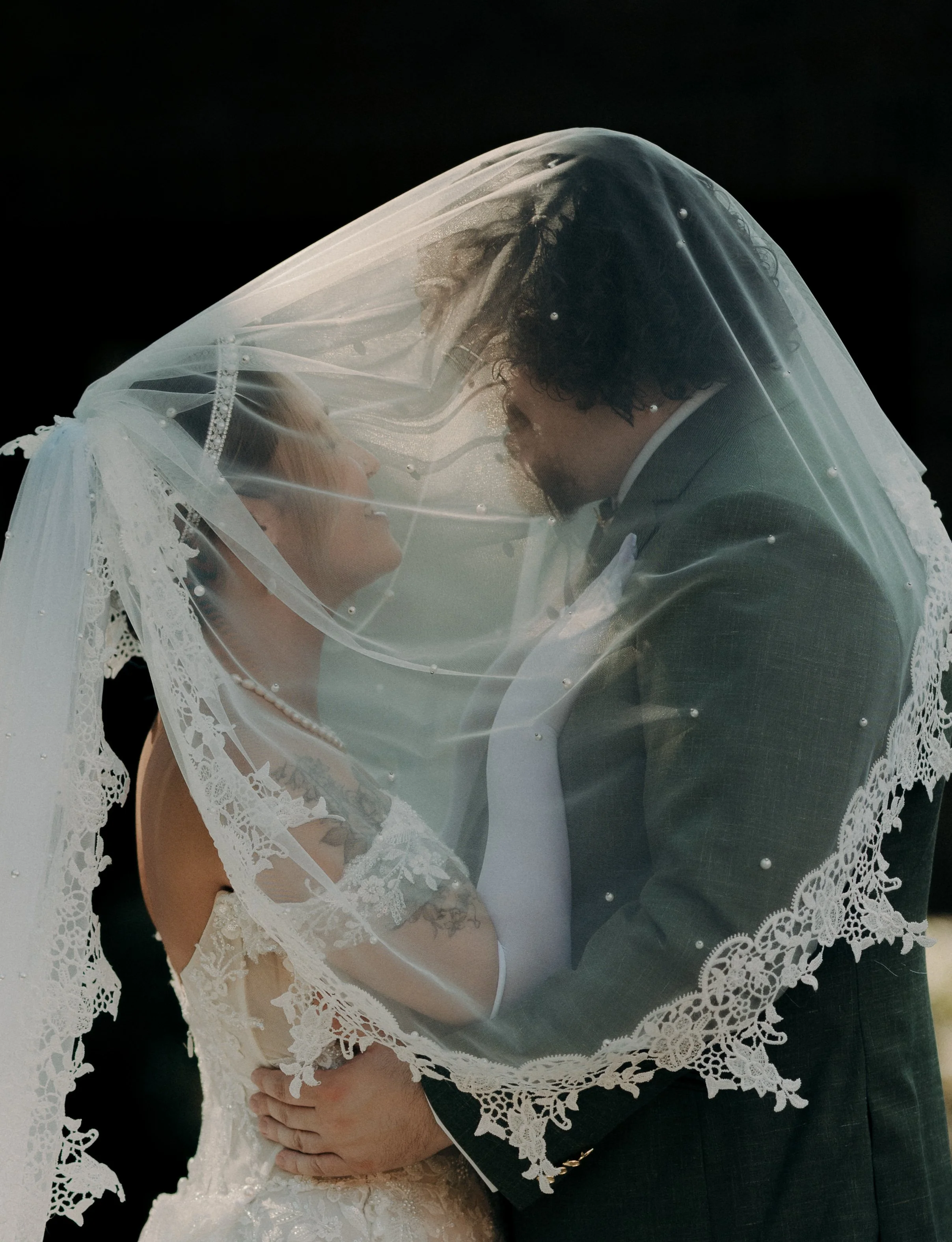 A bride and groom are smiling and looking into each other's eyes under a lace wedding veil during their wedding celebration.
