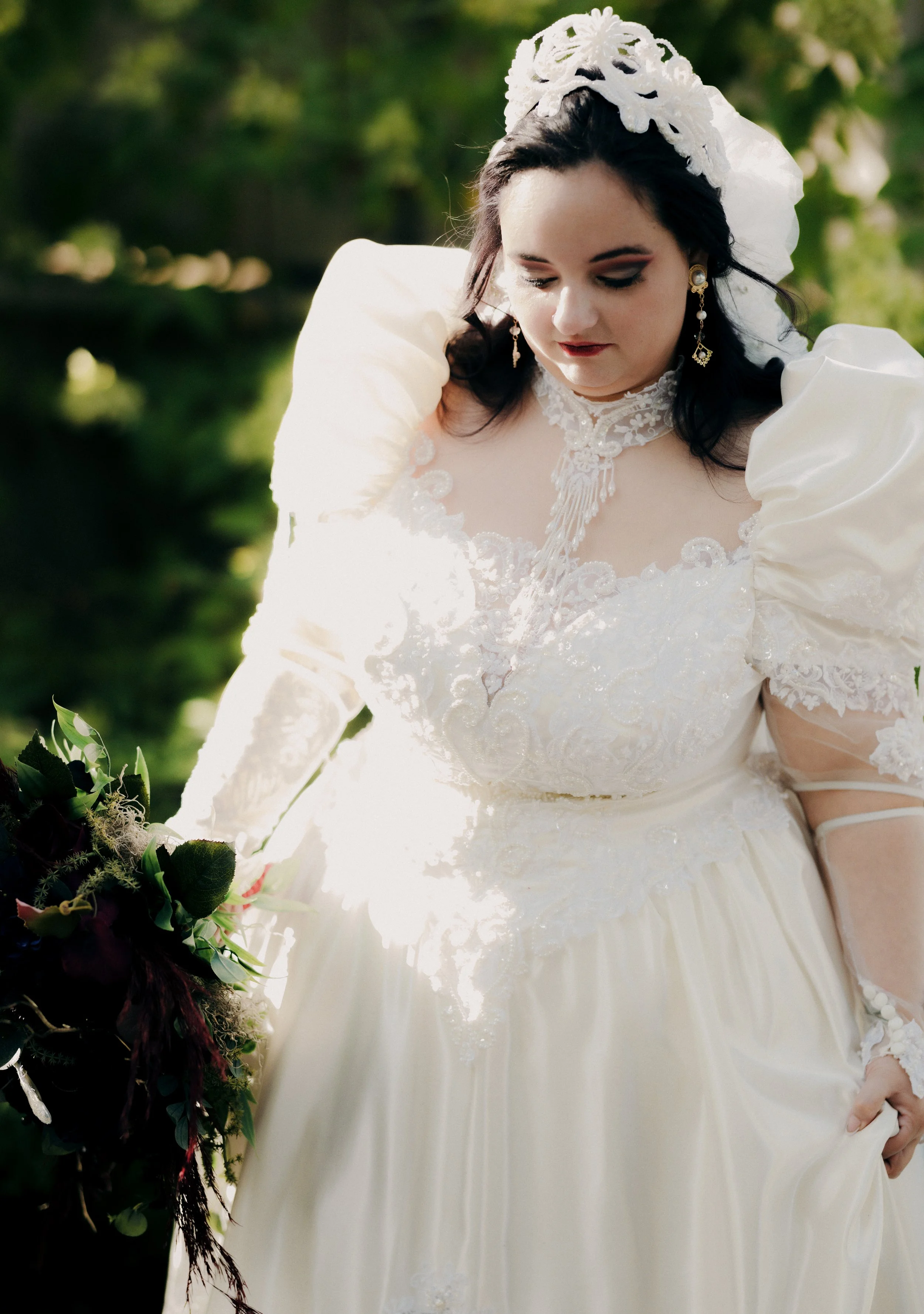 Woman in a vintage white wedding dress with puffy sleeves and lace details, holding a bouquet of dark-colored flowers, outdoors with greenery in the background.