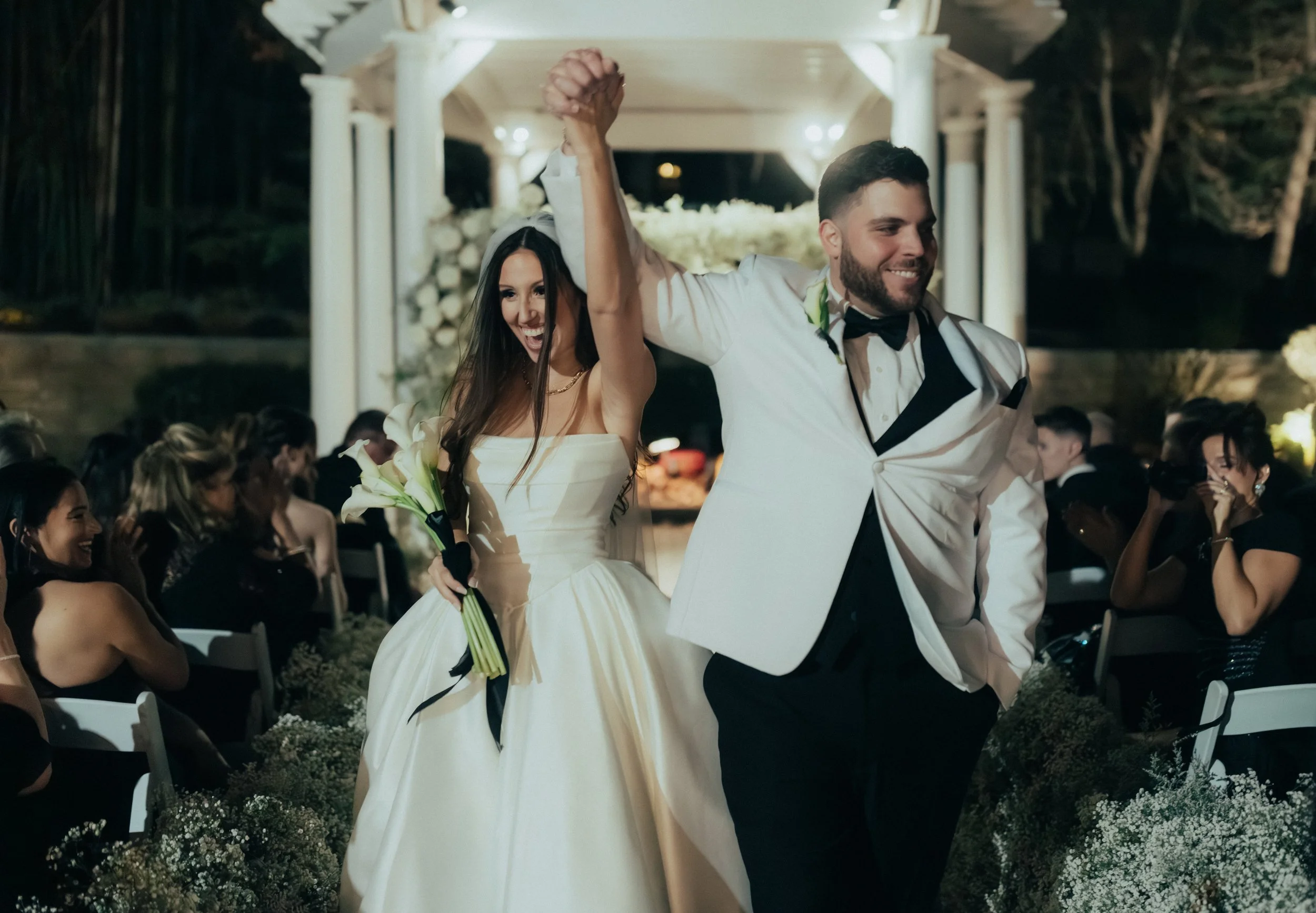 A bride and groom celebrating at their wedding reception, holding hands raised in victory, with guests in the background.