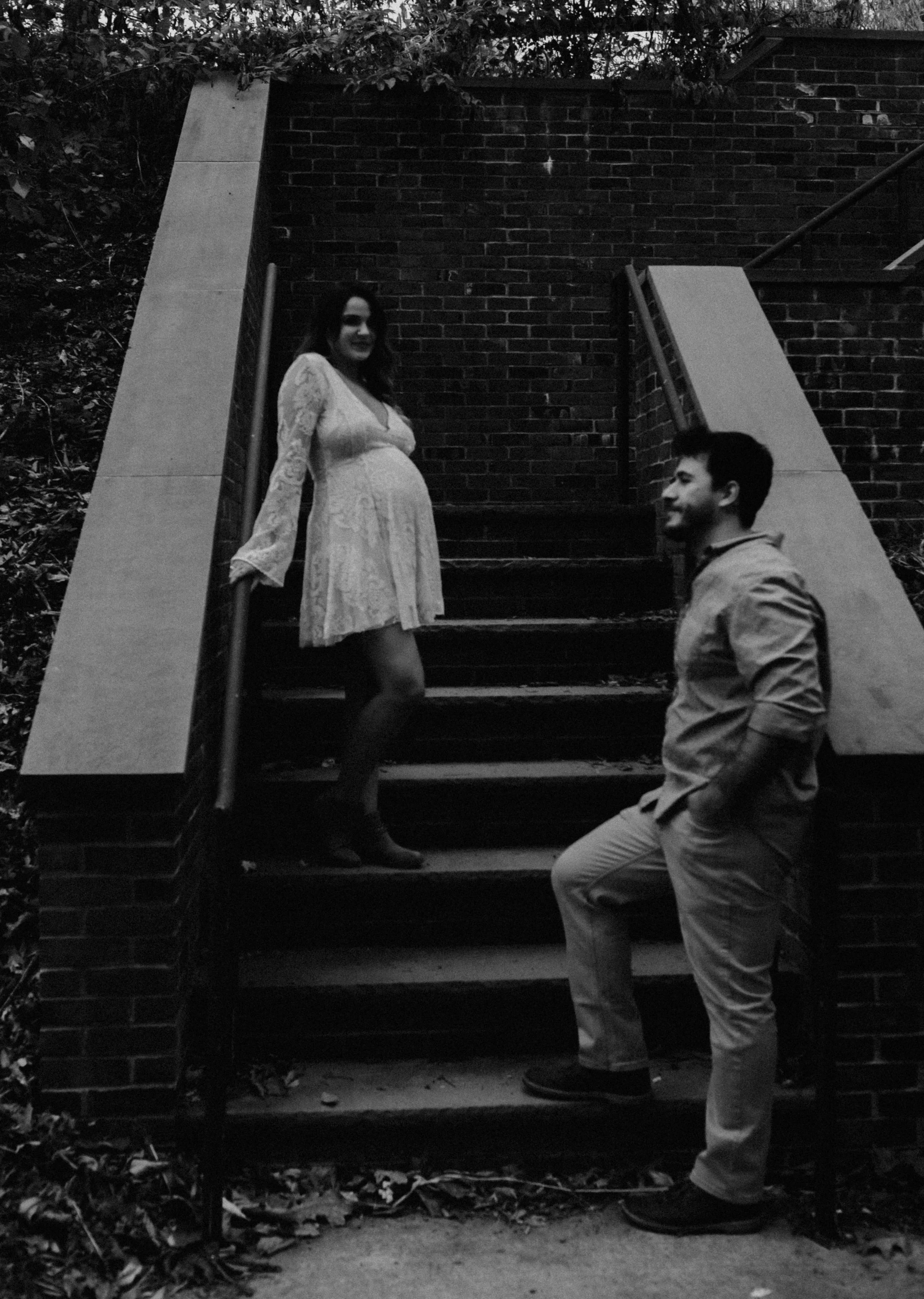 A pregnant woman in a lace dress standing on outdoor stairs, talking to a man in casual clothing, with a brick wall and trees in the background.