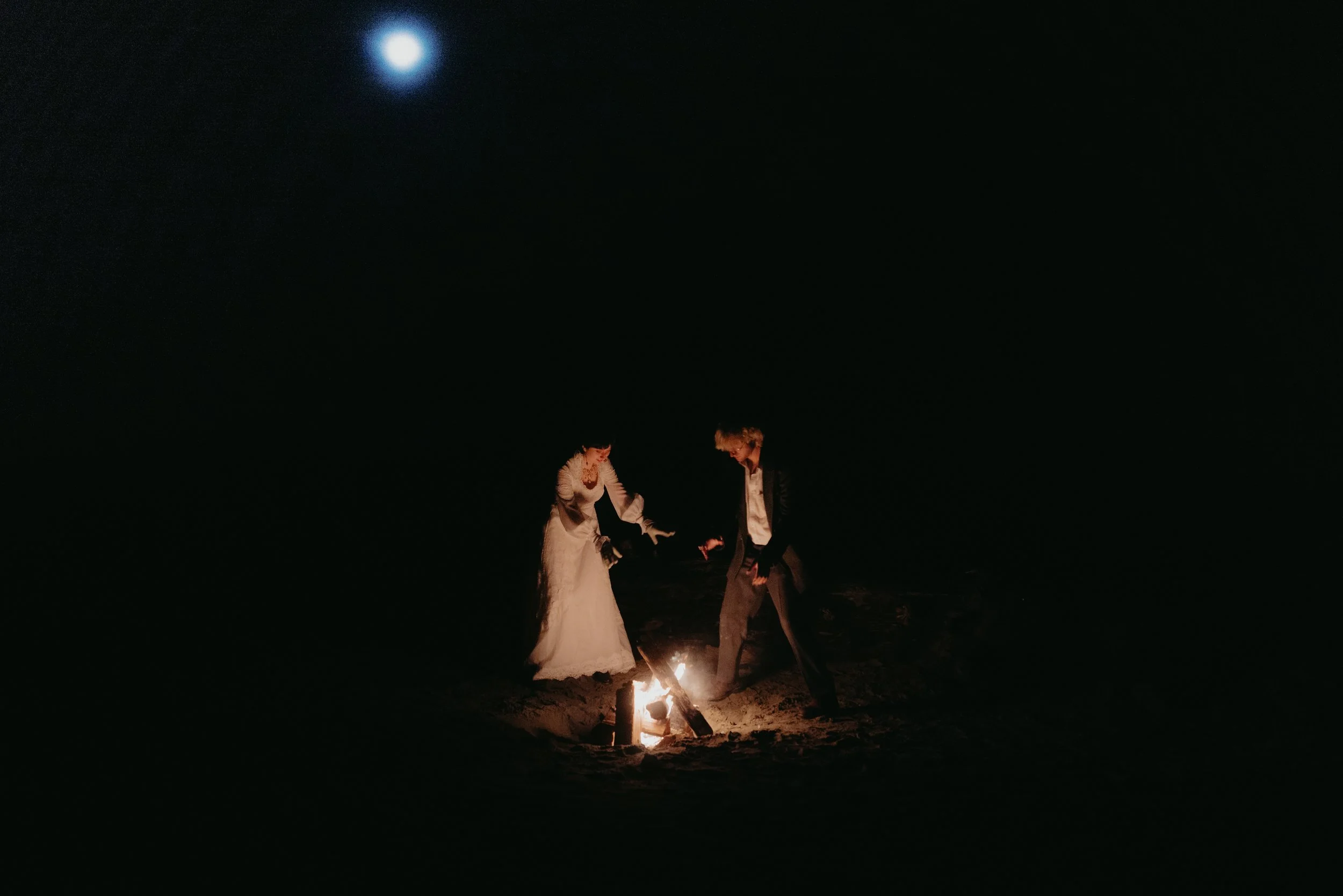 A couple dressed in wedding attire, a bride and groom, standing near a small bonfire on a dark night beach, with the moon visible in the sky.