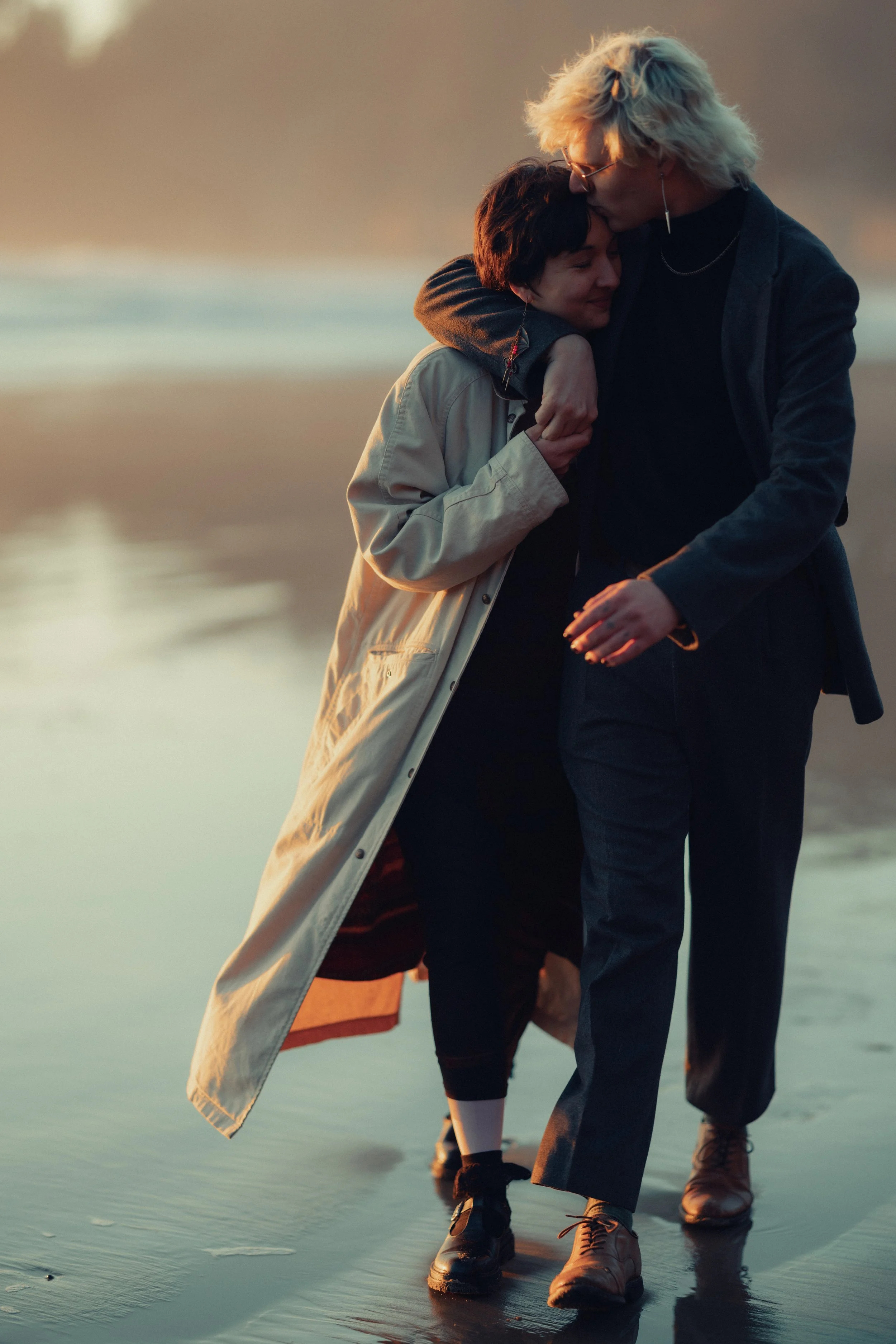 Two people walking on the beach at sunset, sharing an intimate moment with one person kissing the other's head.