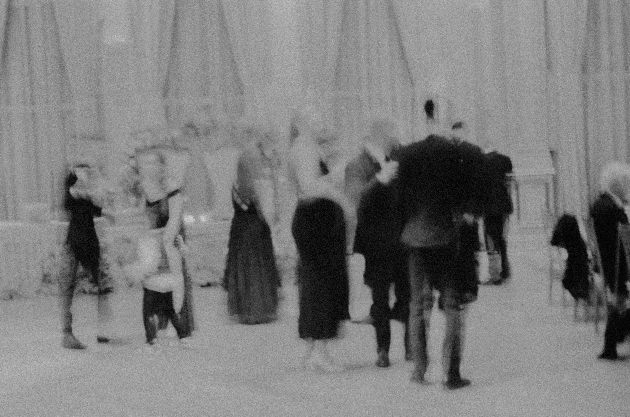 Black and white photo of several people in formal attire dancing in a large room with high ceilings and decorative wall details.