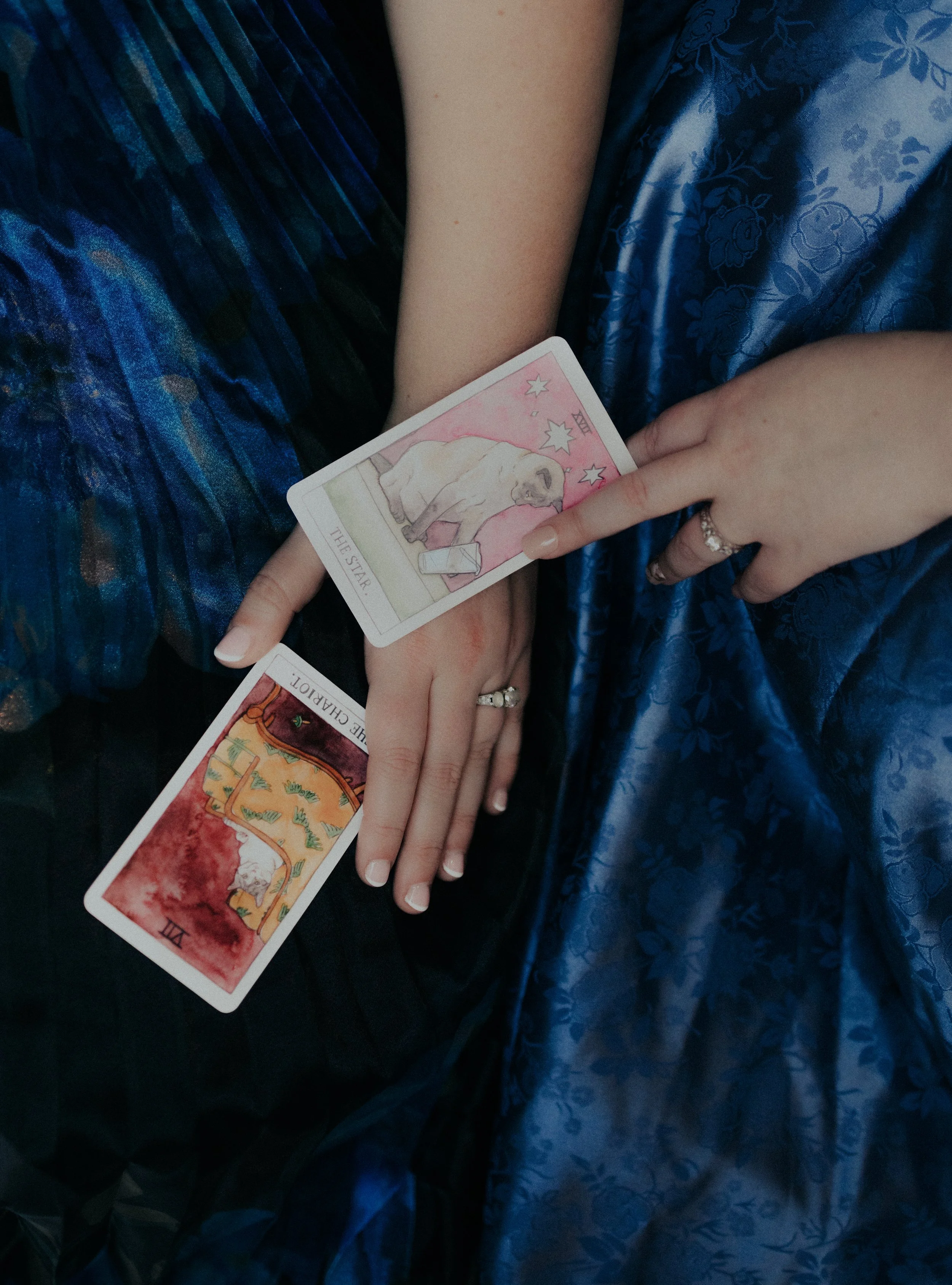 Two people with intertwined hands, each holding tarot cards. One person wears a blue satin dress, and the other wears a multicolored skirt with a pattern. The tarot cards show the Star and The Chariot.