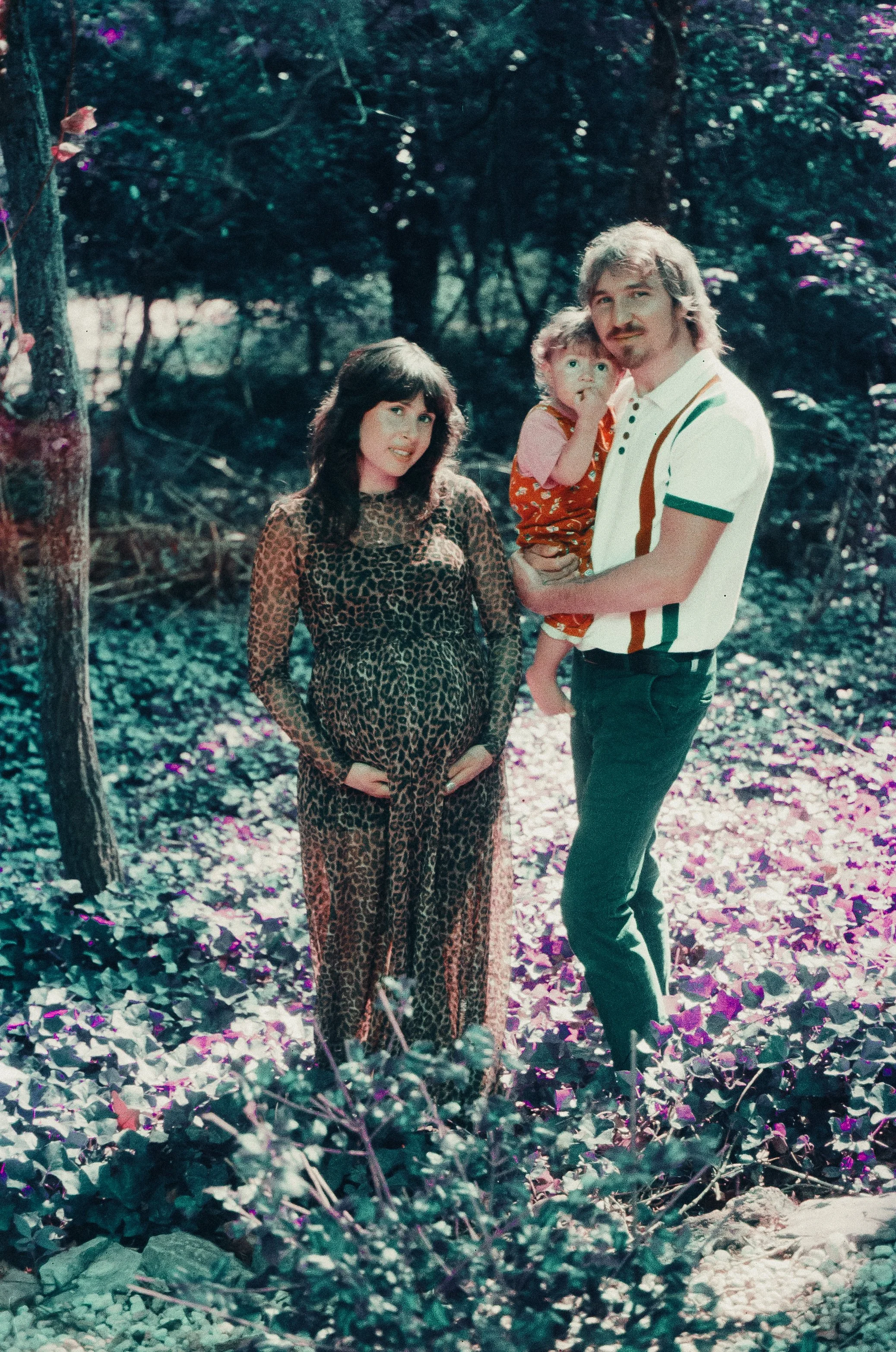 A family of three in a wooded area. The woman is wearing a leopard-print long dress, pregnant, with dark hair. The man with light-colored hair and goatee is holding a child and wearing a white polo shirt with colored stripes and dark pants. The child