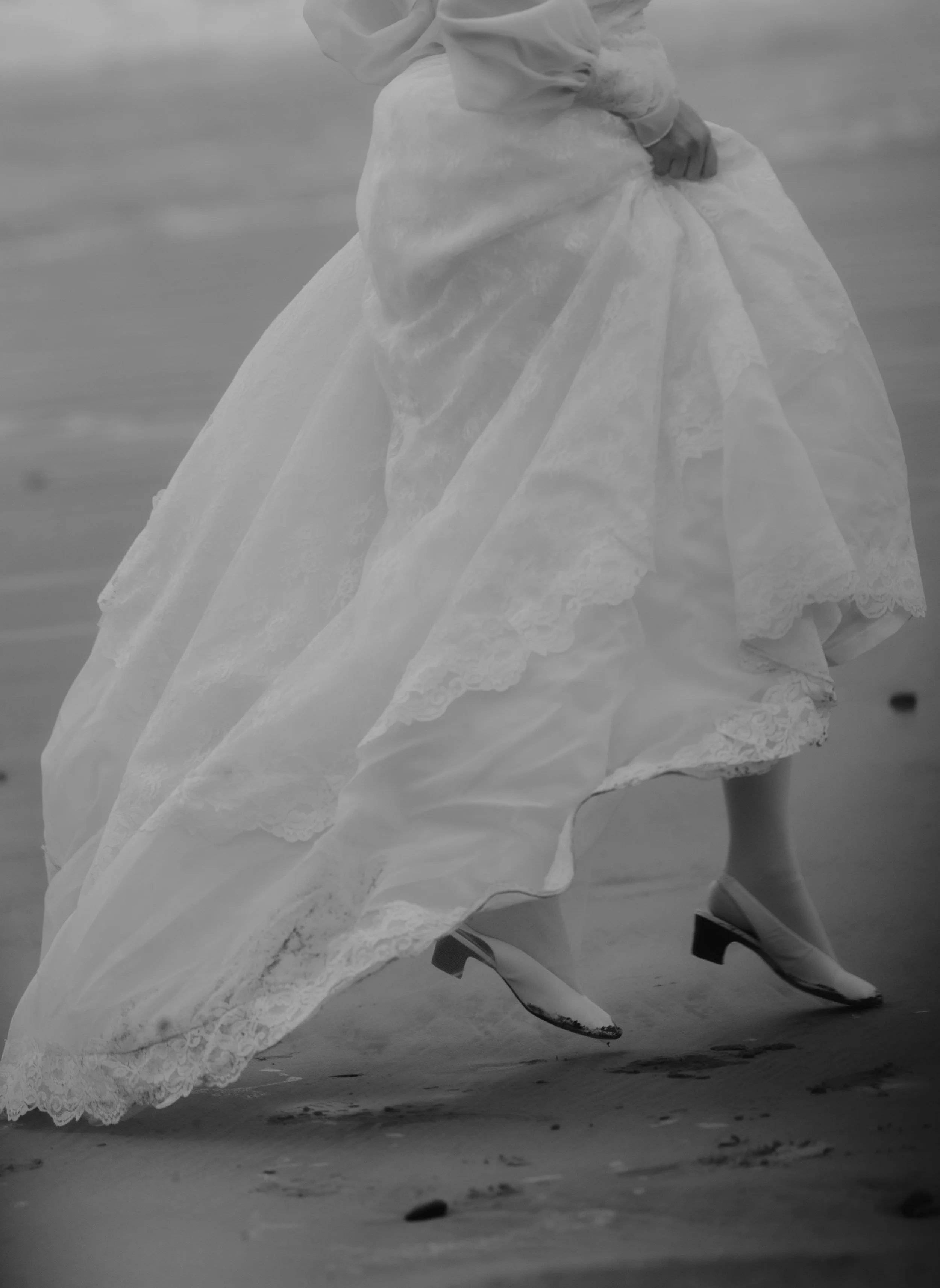 A person wearing a vintage wedding dress and high heels walking on the beach, with the dress's lace and fabric flowing as they walk.