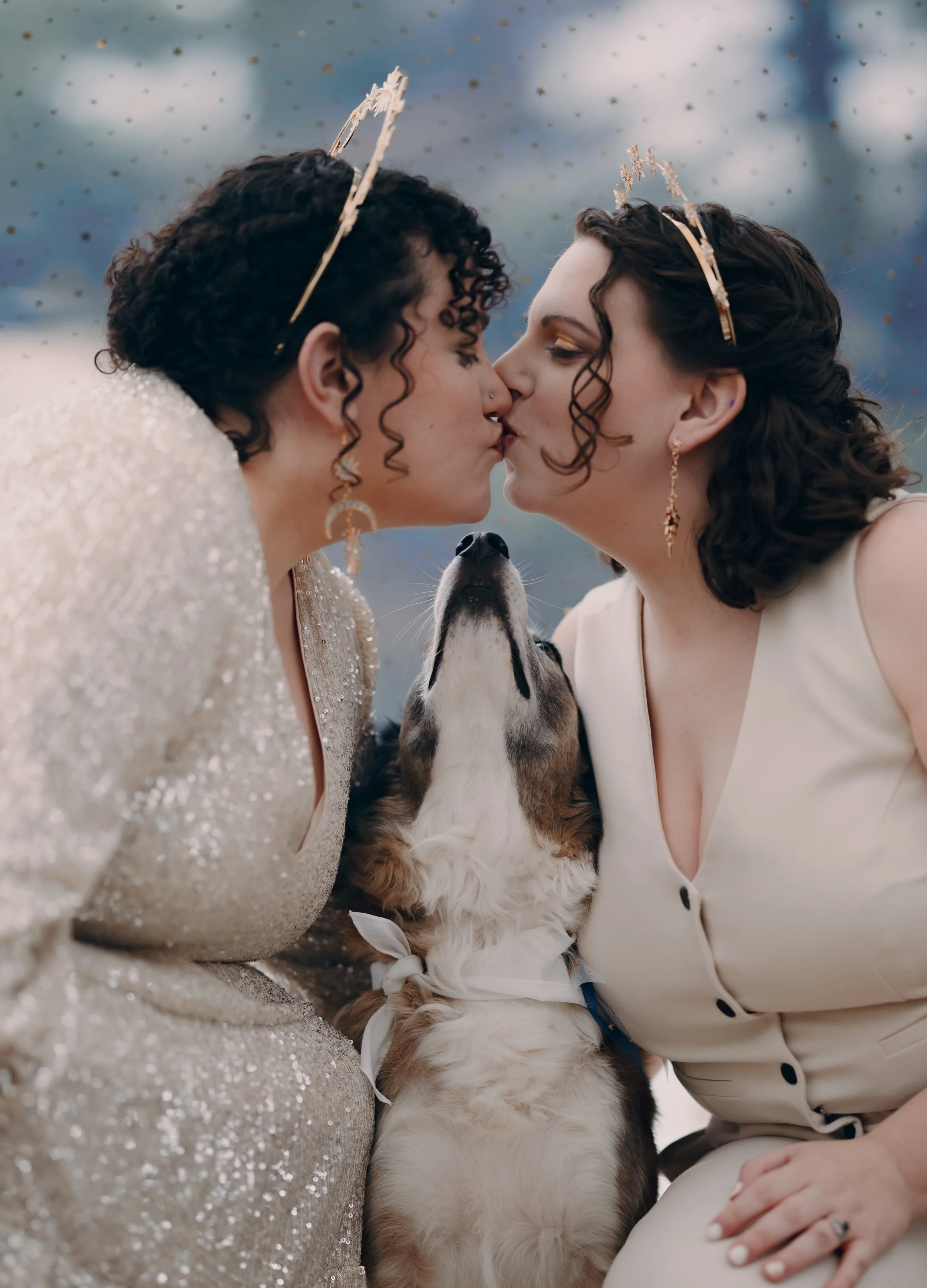 Two women wearing tiaras and earrings are kissing each other while a dog between them is licking one woman's face during a wedding celebration.