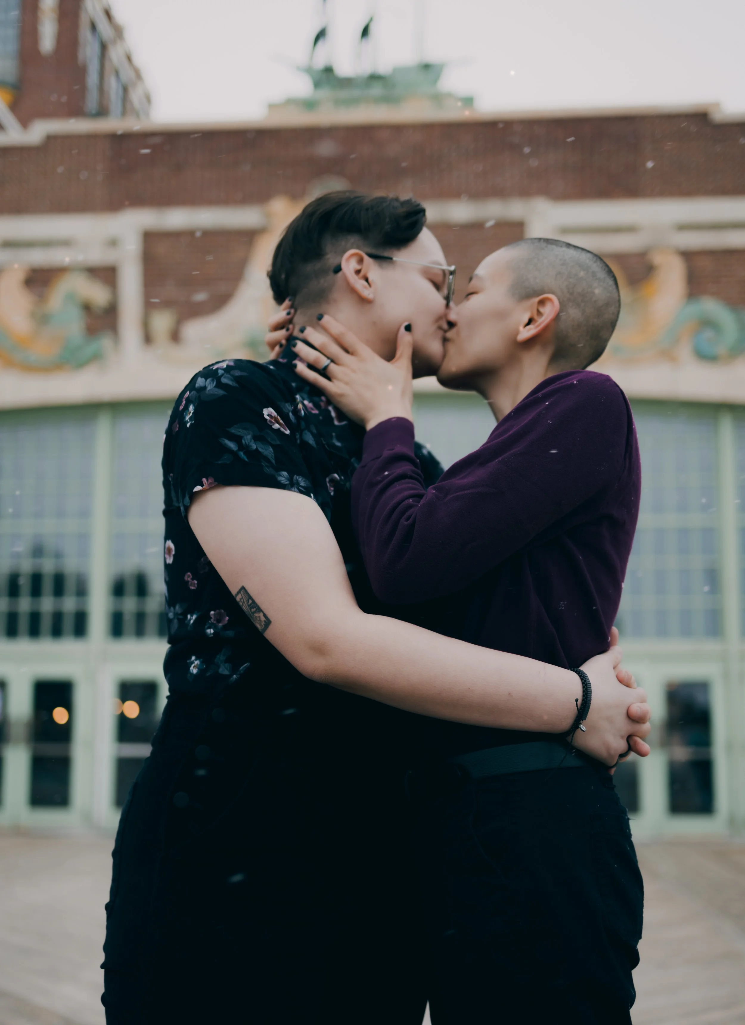 Two women sharing a kiss outdoors in front of decorative building architecture.