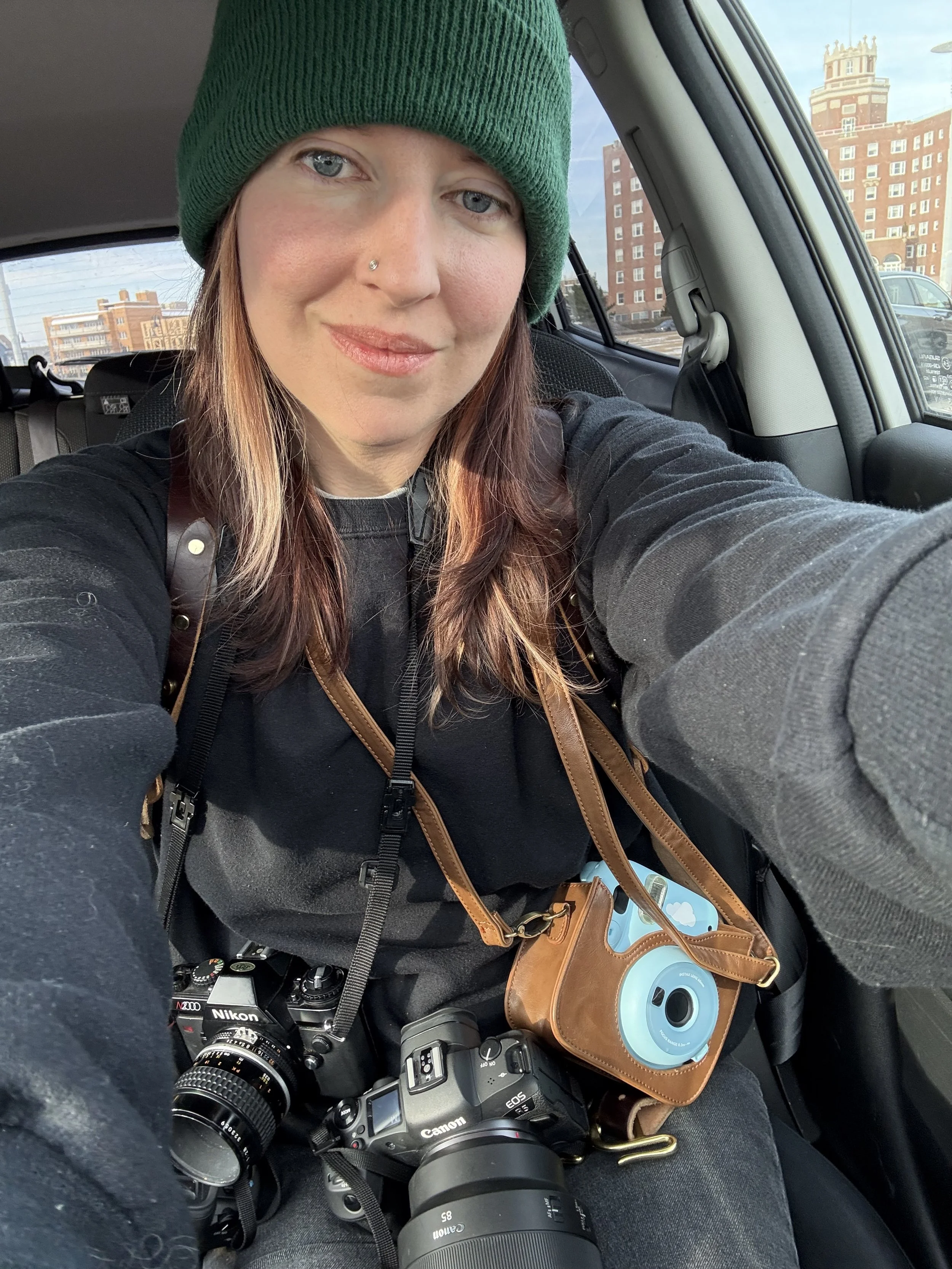 A woman wearing a green beanie and black sweatshirt taking a selfie inside a car. She has a nose piercing and is carrying two cameras around her neck, one Nikon and one Canon, along with a brown camera case with a blue cloud design.