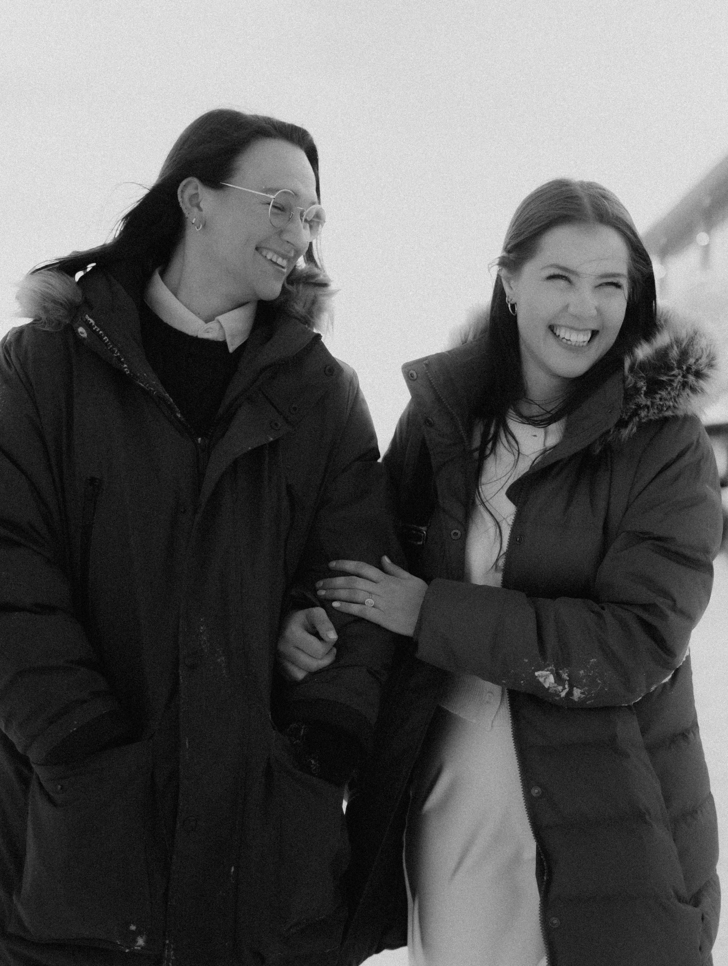 Two women wearing winter jackets laughing and smiling outdoors in black and white.