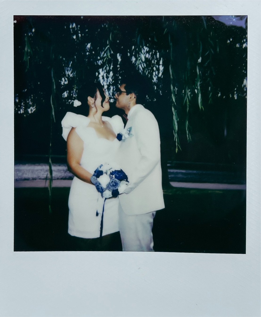 A bride and groom sharing a kiss outdoors, with green hanging trees in the background, during a wedding ceremony.