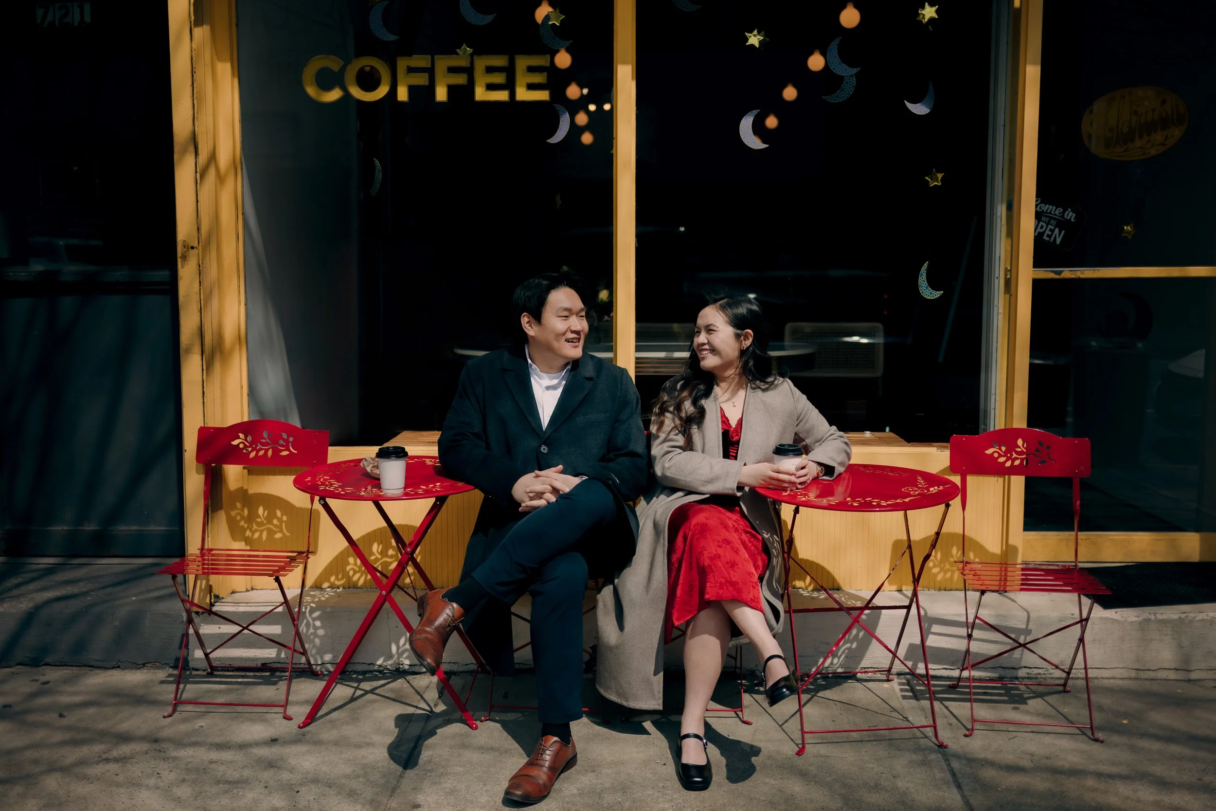 A man and woman sitting at a red outdoor cafe table in front of a coffee shop, smiling and looking at each other, each holding a coffee cup, with additional empty chairs around them.