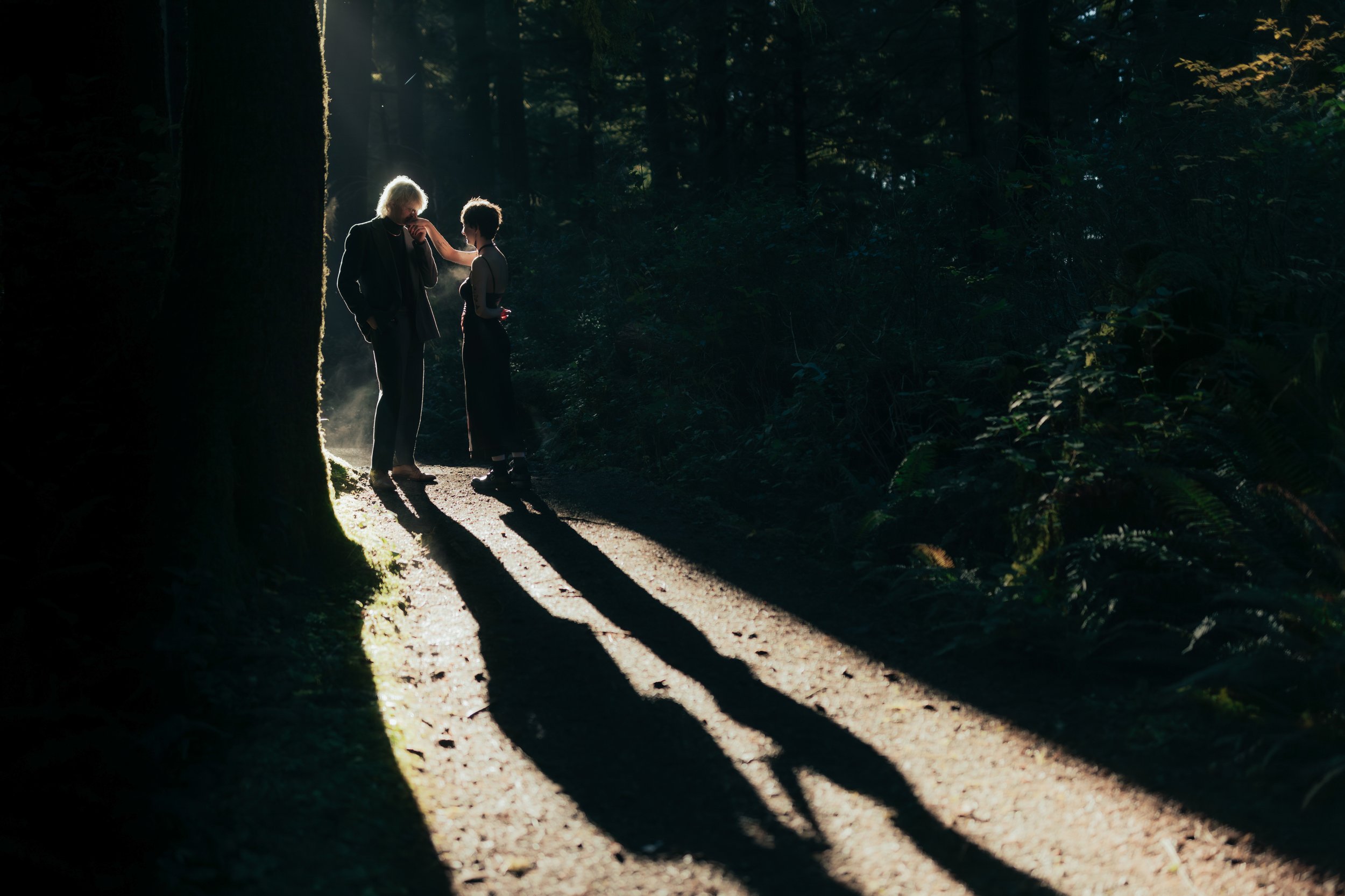 Two people stand on a sunlit forest trail, with long shadows cast by trees, engaging in a conversation with one touching the other's face.