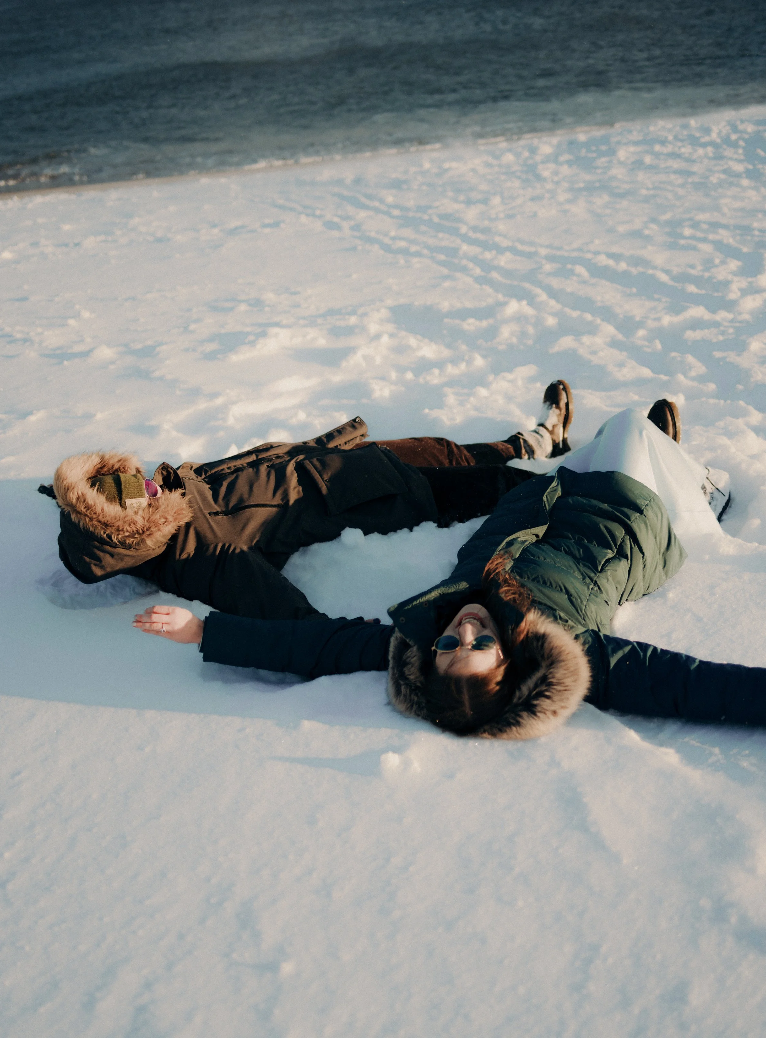 Two women lying on snow-covered ground in winter clothing, wearing fur-lined hoods and sunglasses, near a body of water.