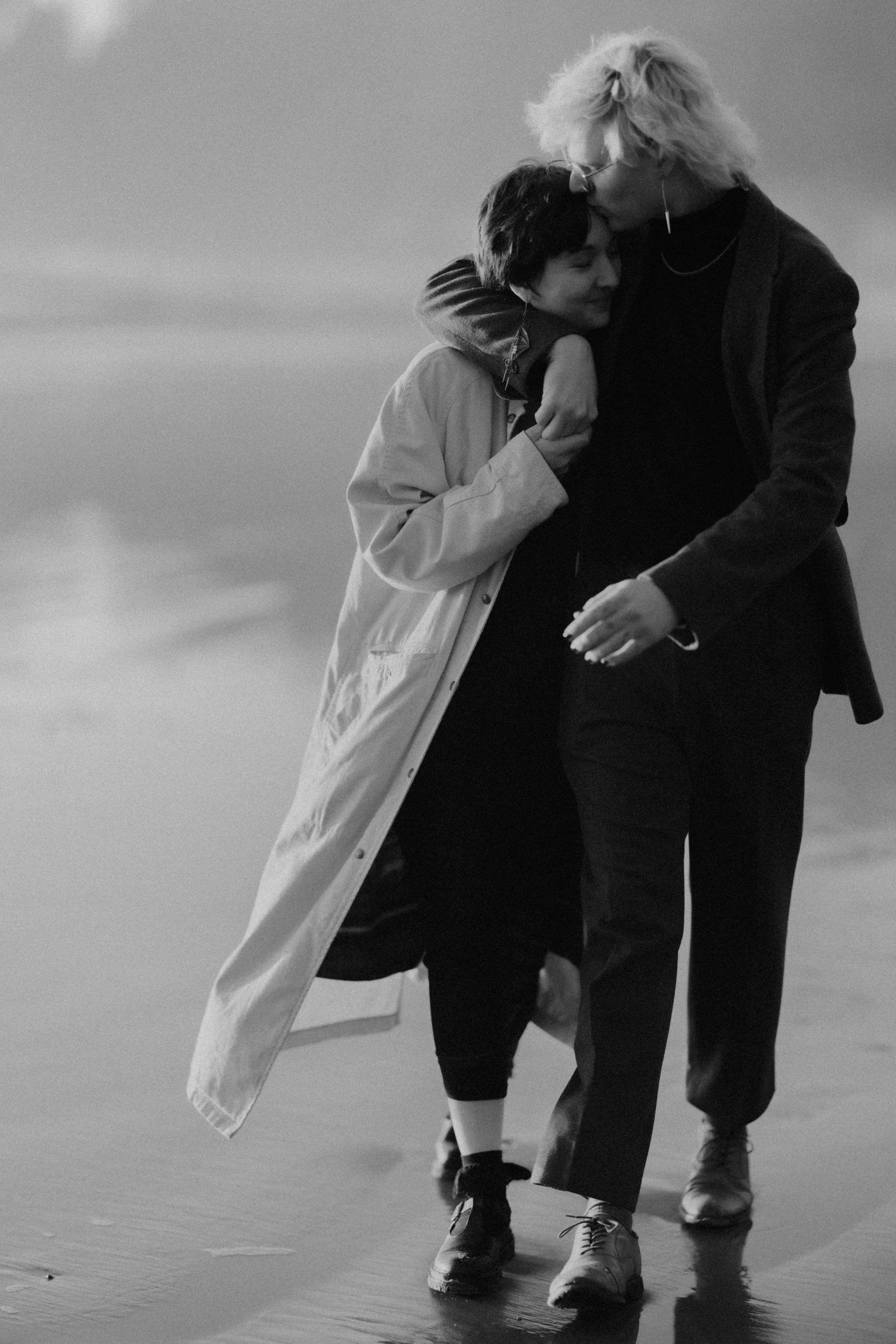 Two women walking closely on a beach, one is kissing the other's forehead, both smiling gently.
